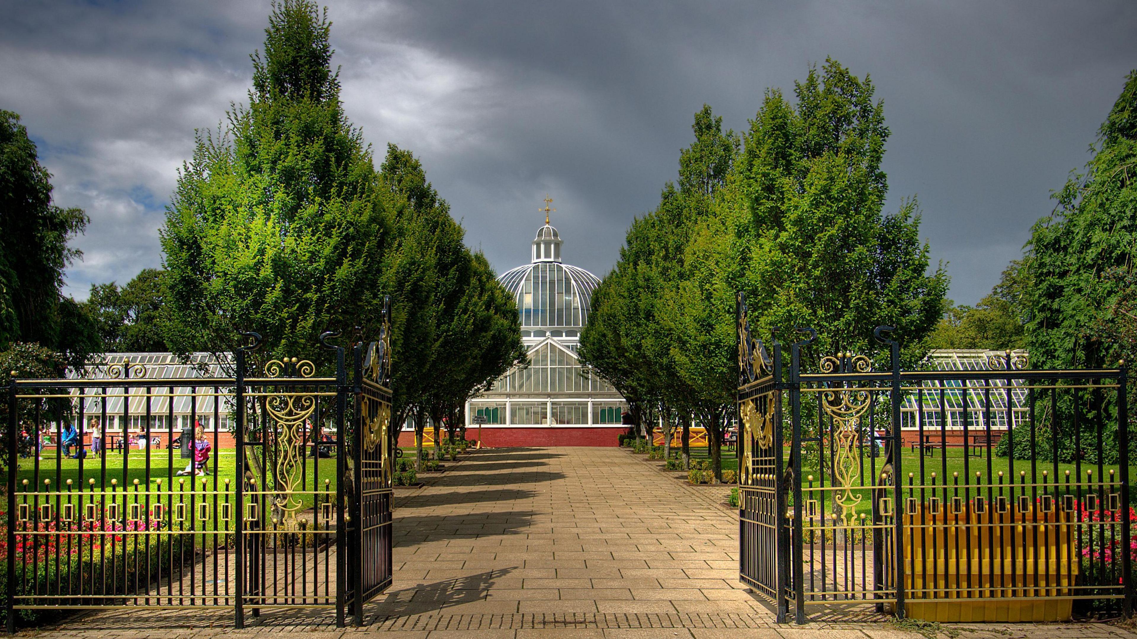 Black gates are open leading to a path up a large glasshouse building. People are playing on the grass outside while dark skies are overhead.