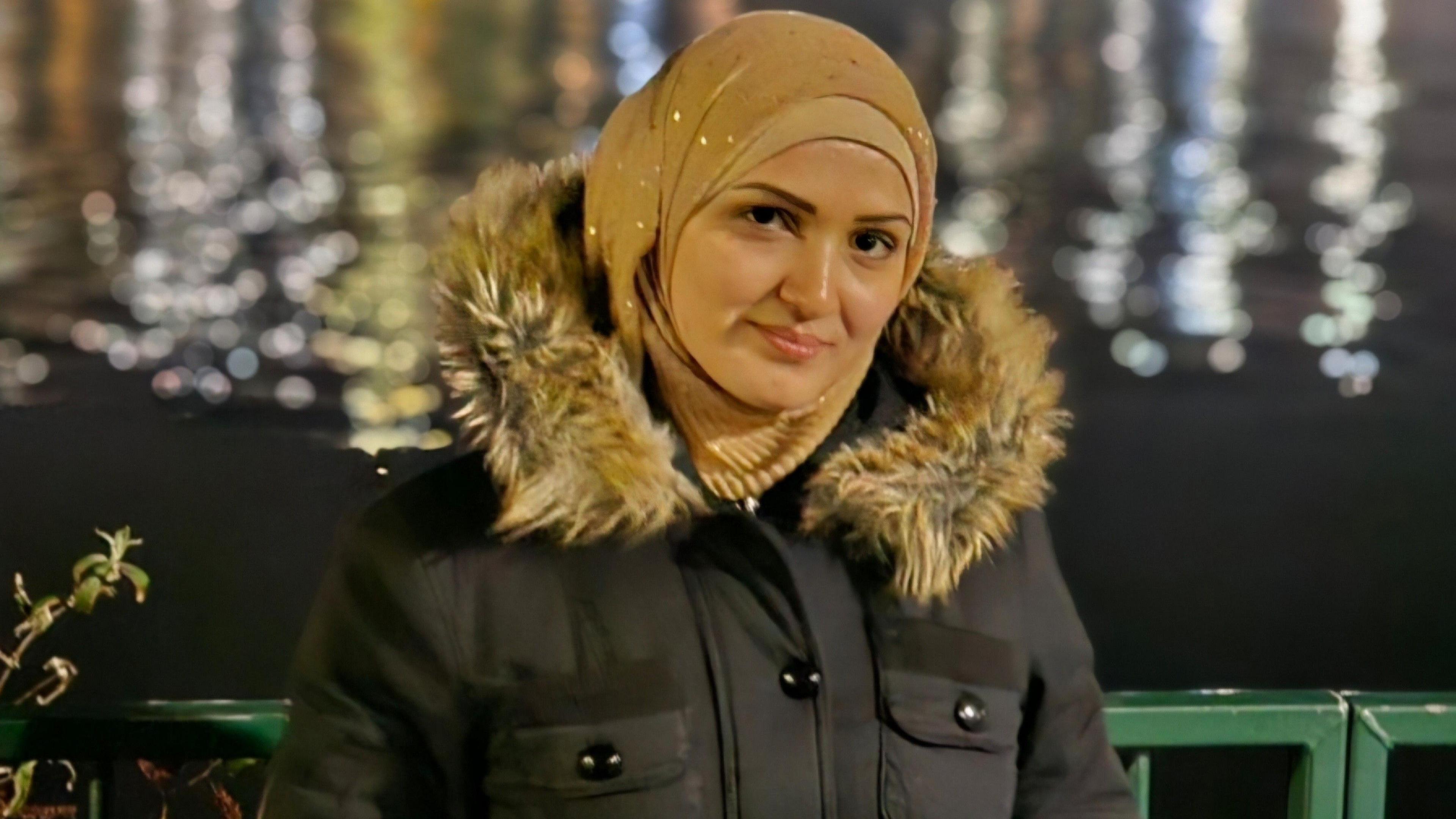 A woman in a gold hijab smiling into the camera. She is wearing a coat with a fur hood and standing in front of a body of water.