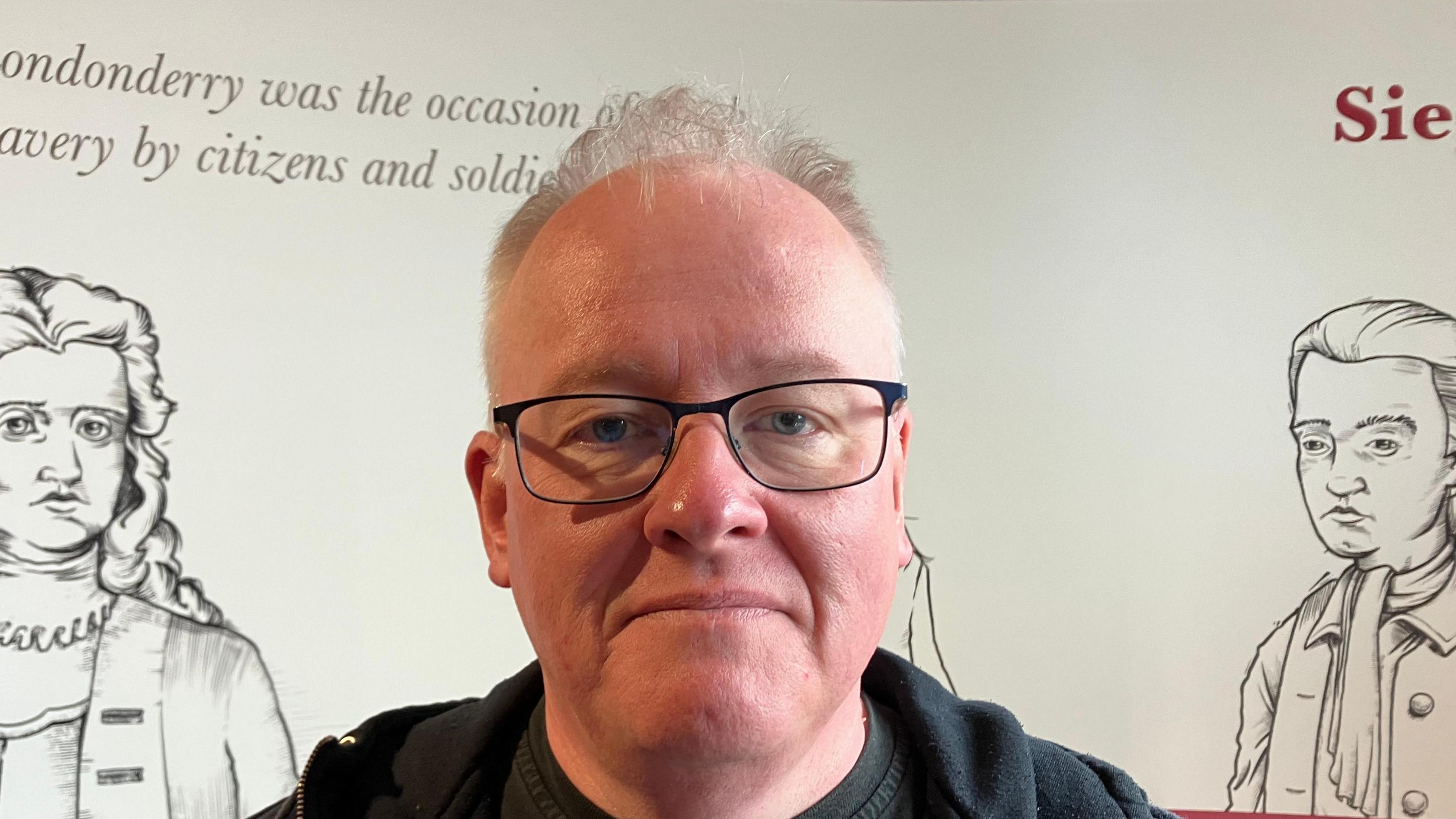 Jonathan Burgess is in a black hoodie and wearing glasses.
He has short hair and is standing behind a white board with information and historical figures on it.