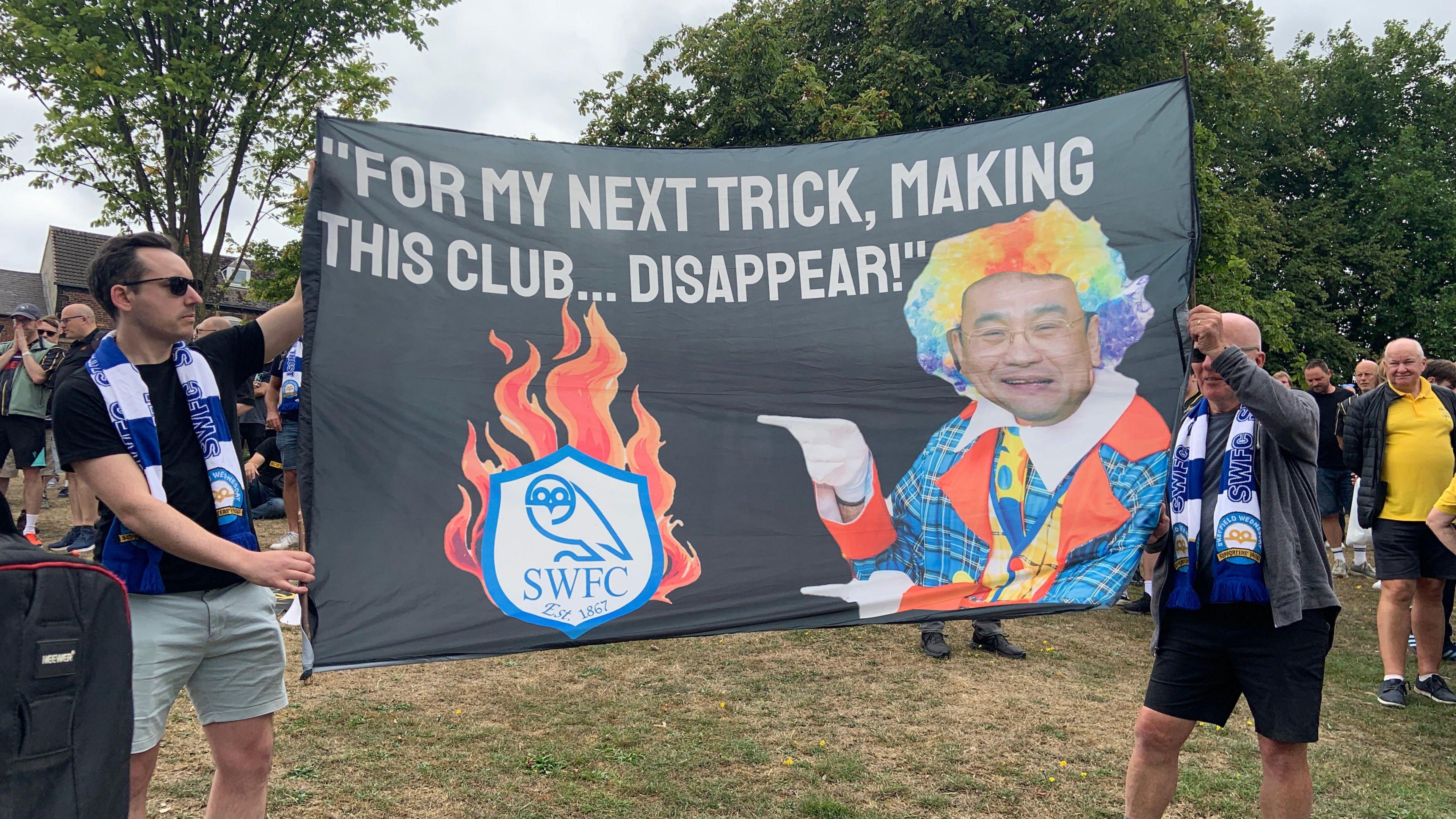 Two men wearing blue and white striped Sheffield Wednesday scarves are holding a large banner. It has an image of a clown and says "for my next trick, making this club disappear"