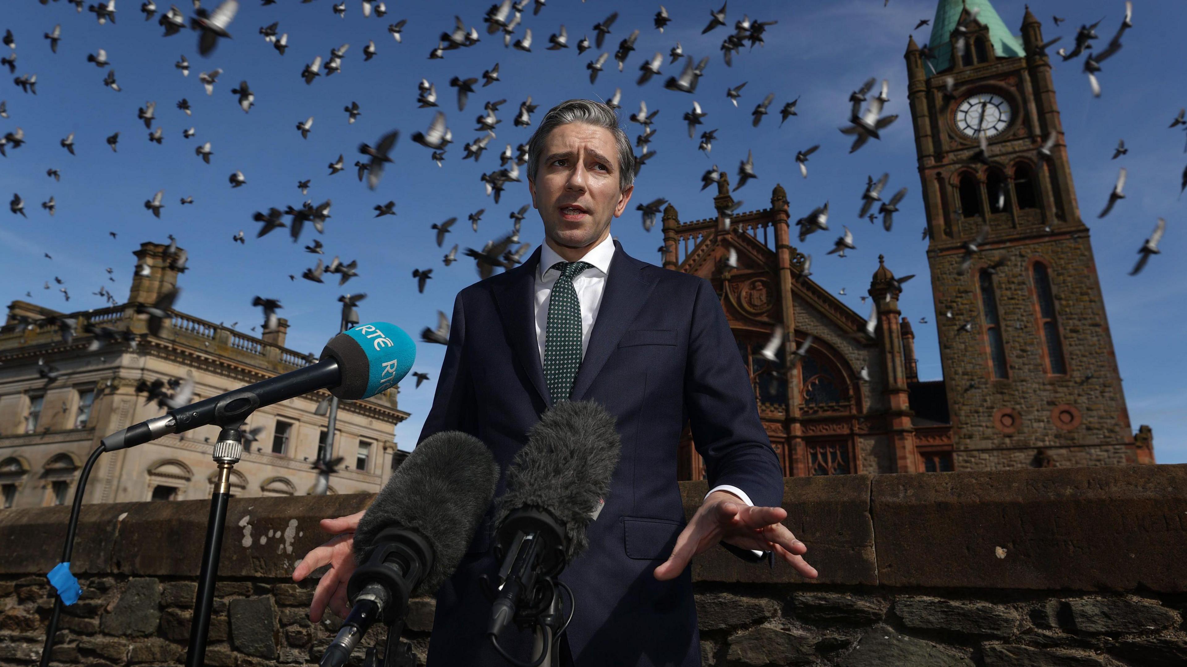 A suited Simon Harris stands on Derrys Walls with the guidlhall behind him. A flock of pigeons flies overhead.