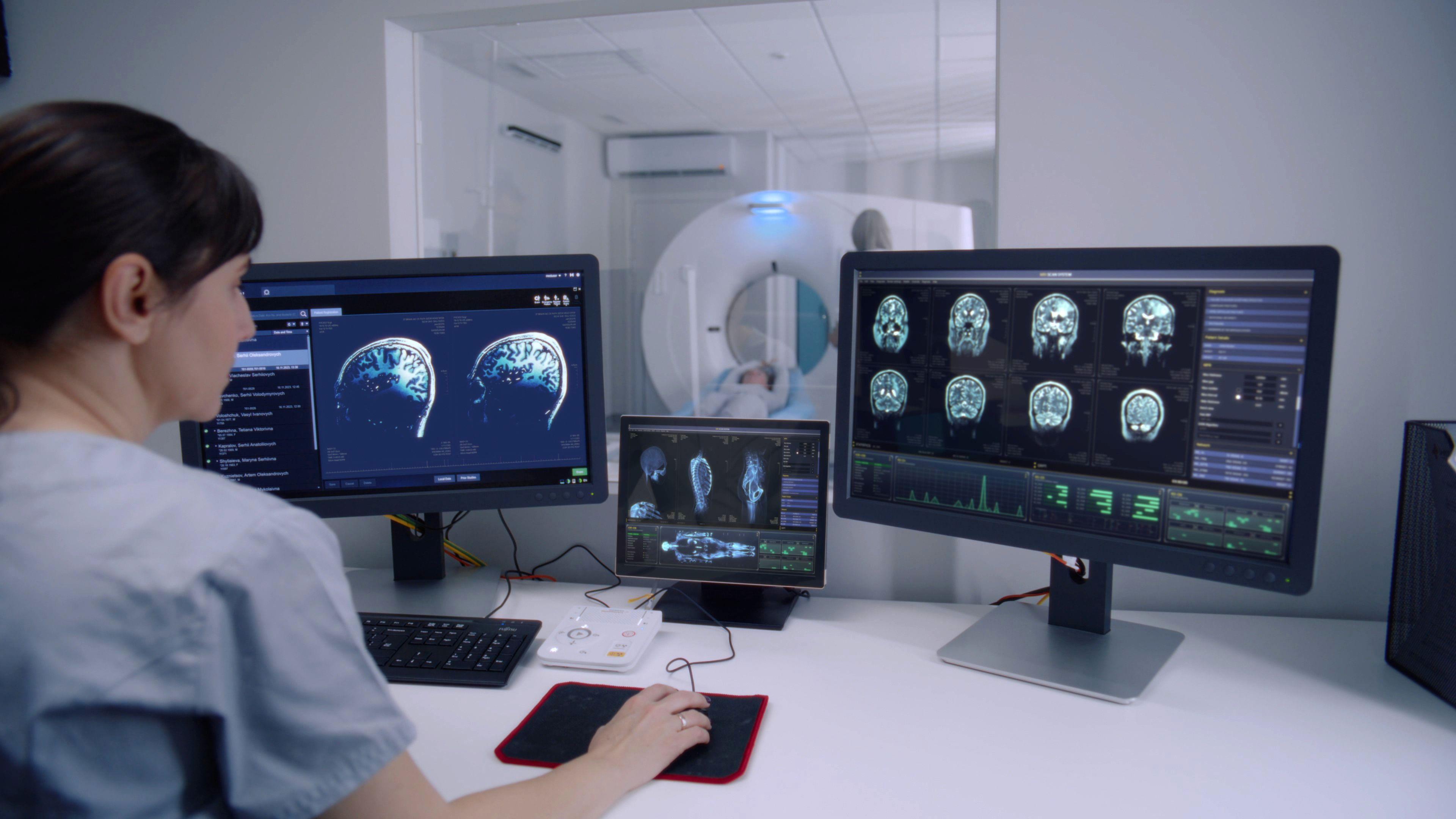 A woman in scrubs sits in front of two computer screens showing images of a person's brain. Behind the computer screens is a window through which we can see a person lying on their back in a CT scanner. 