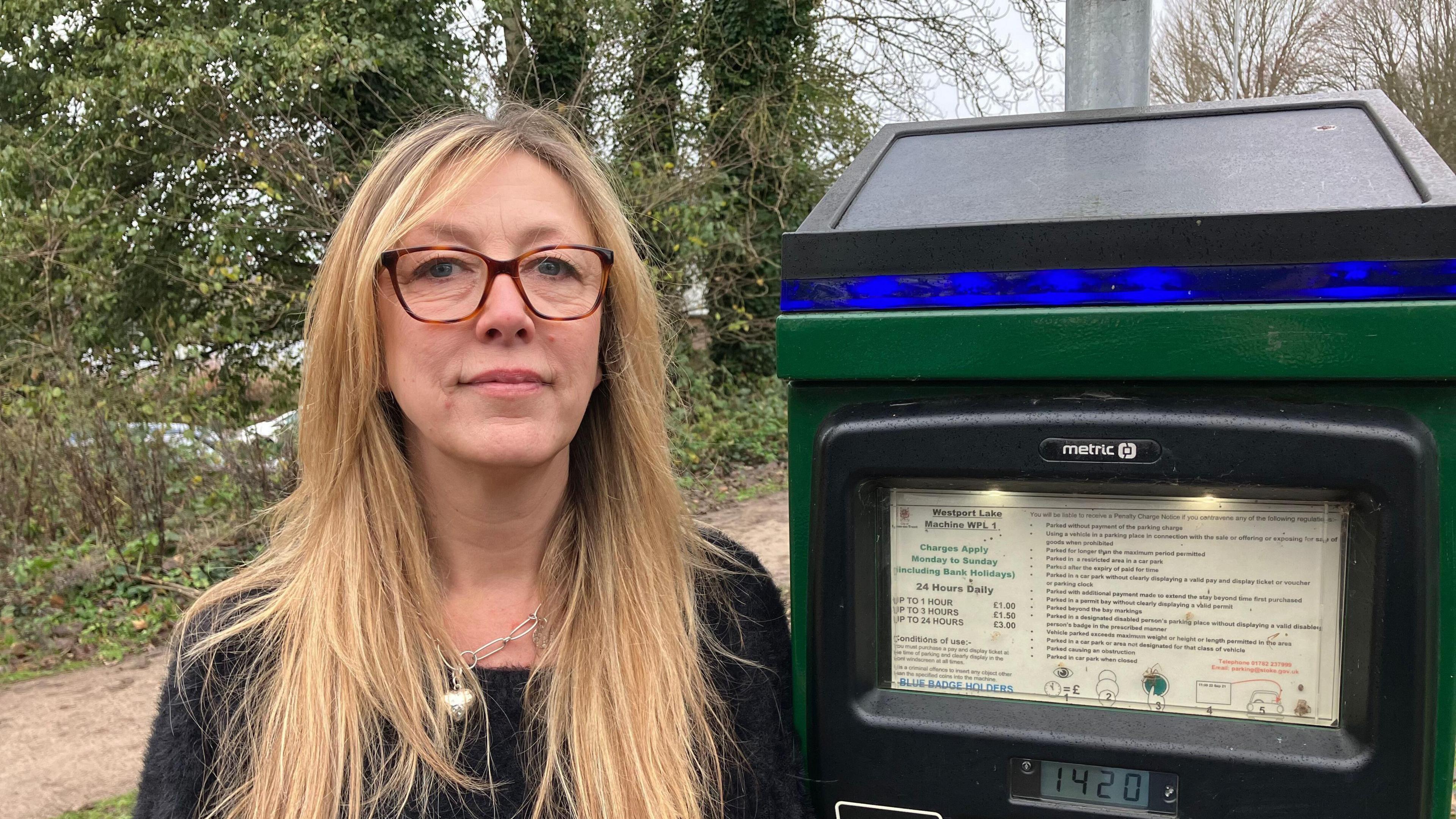 A women with brunet hair, wearing brown glasses and a black jumper. She is stood next to a green parking meter.