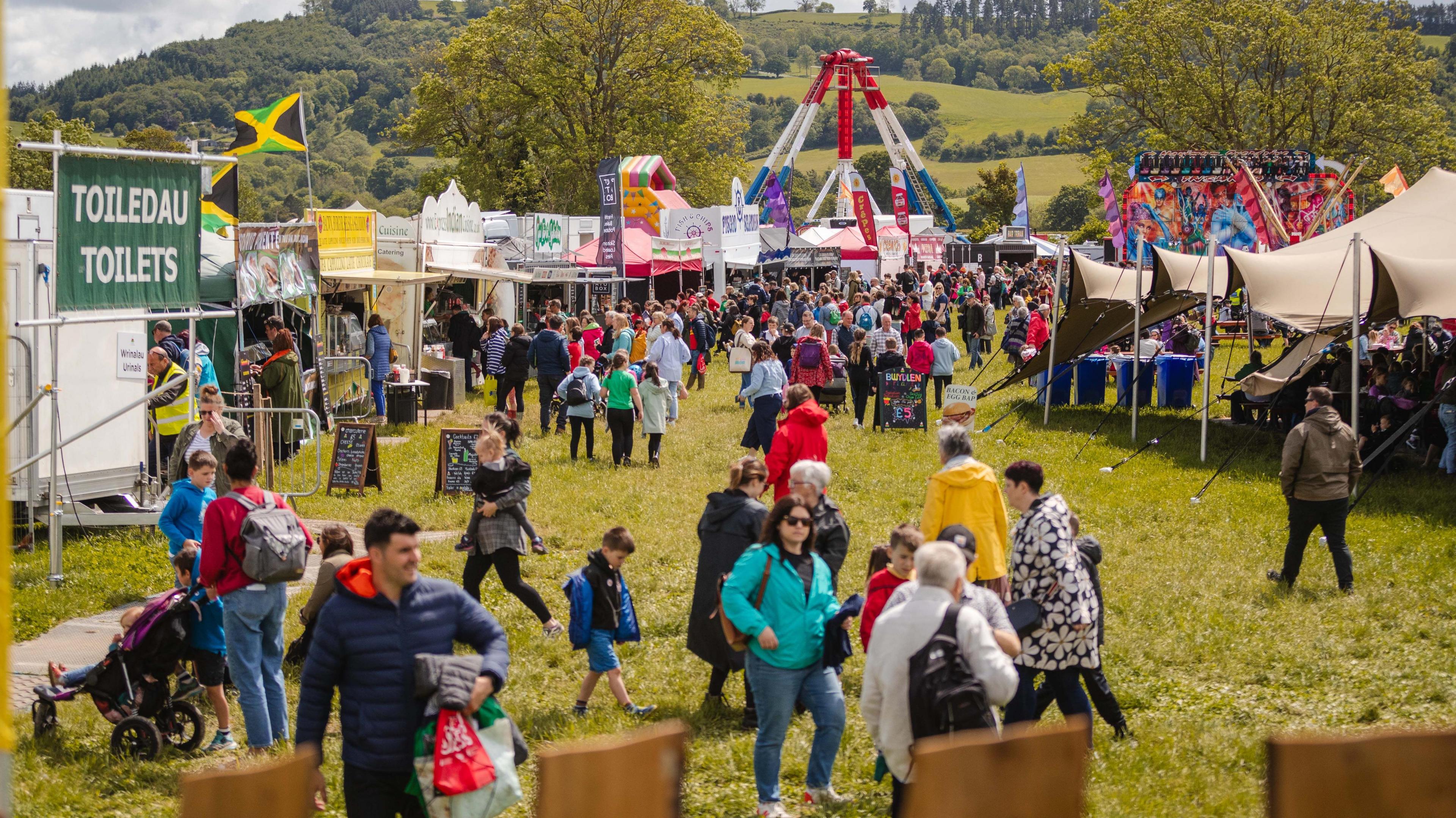 The festival field, people walk around the rides and stalls.