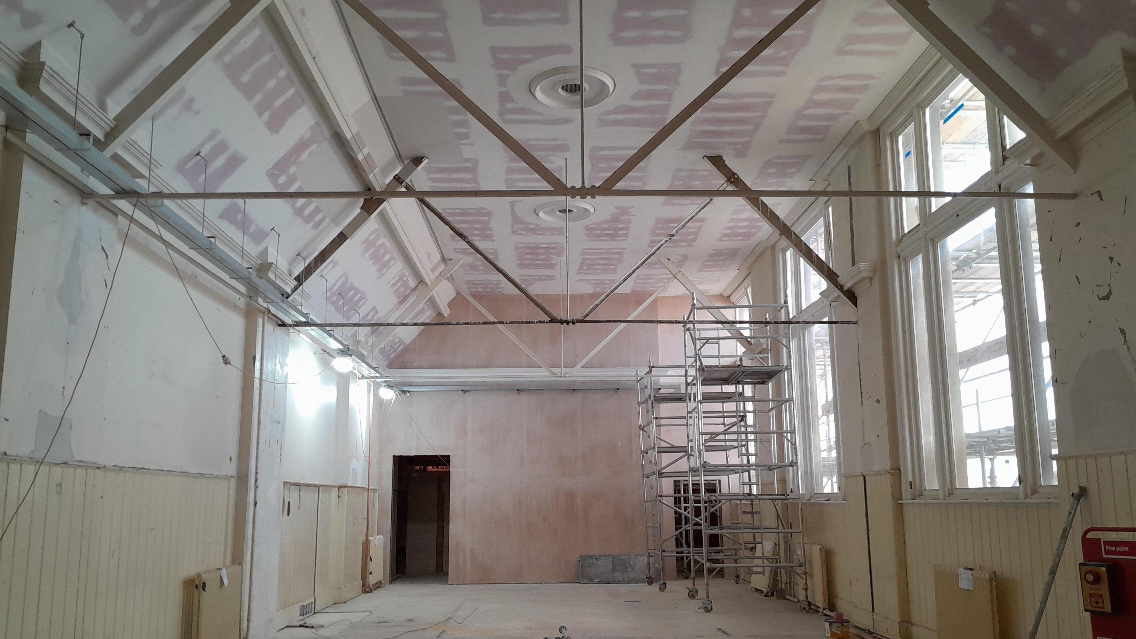 View of a room with a high ceiling which is painted pink and white. The walls of the room are yellow and white, and there are two scaffold towers, made of silver metal, against the right wall. 