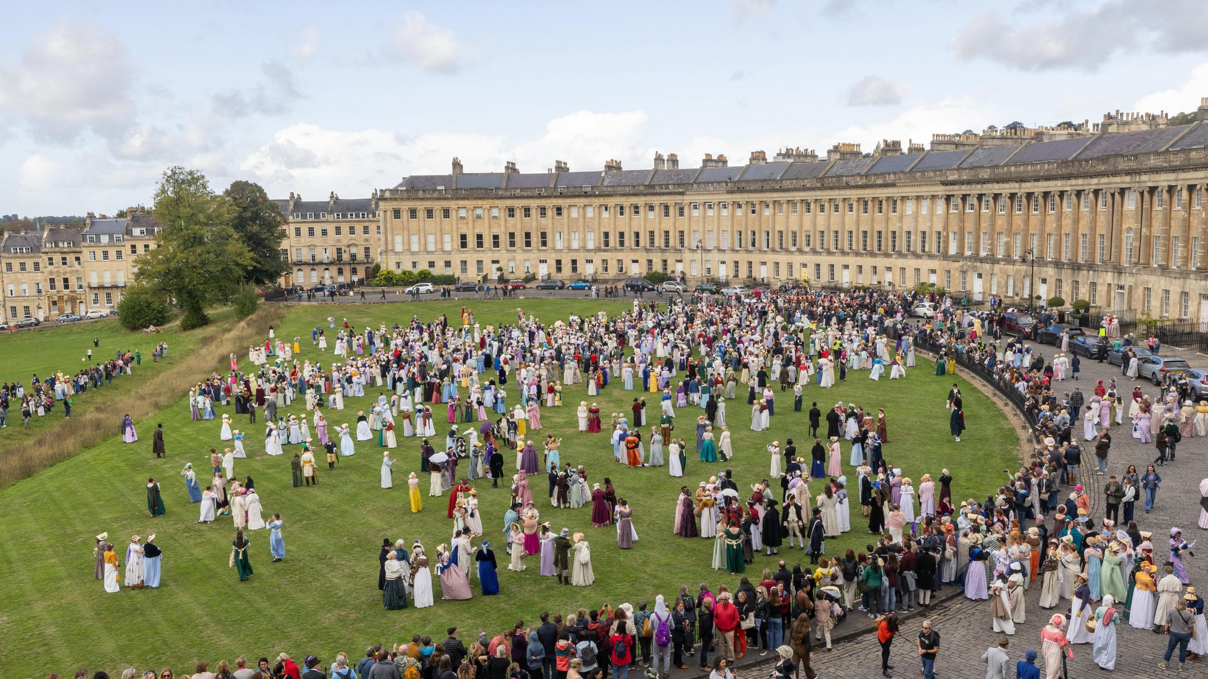 A large crowd of people wearing regency costumes for the Jane Austen Festival in Bath. They are pictured on the grass in front of the Royal Crescent, a historic, semi circle shaped building made out of Cotswold stone. The property has many windows on it. 