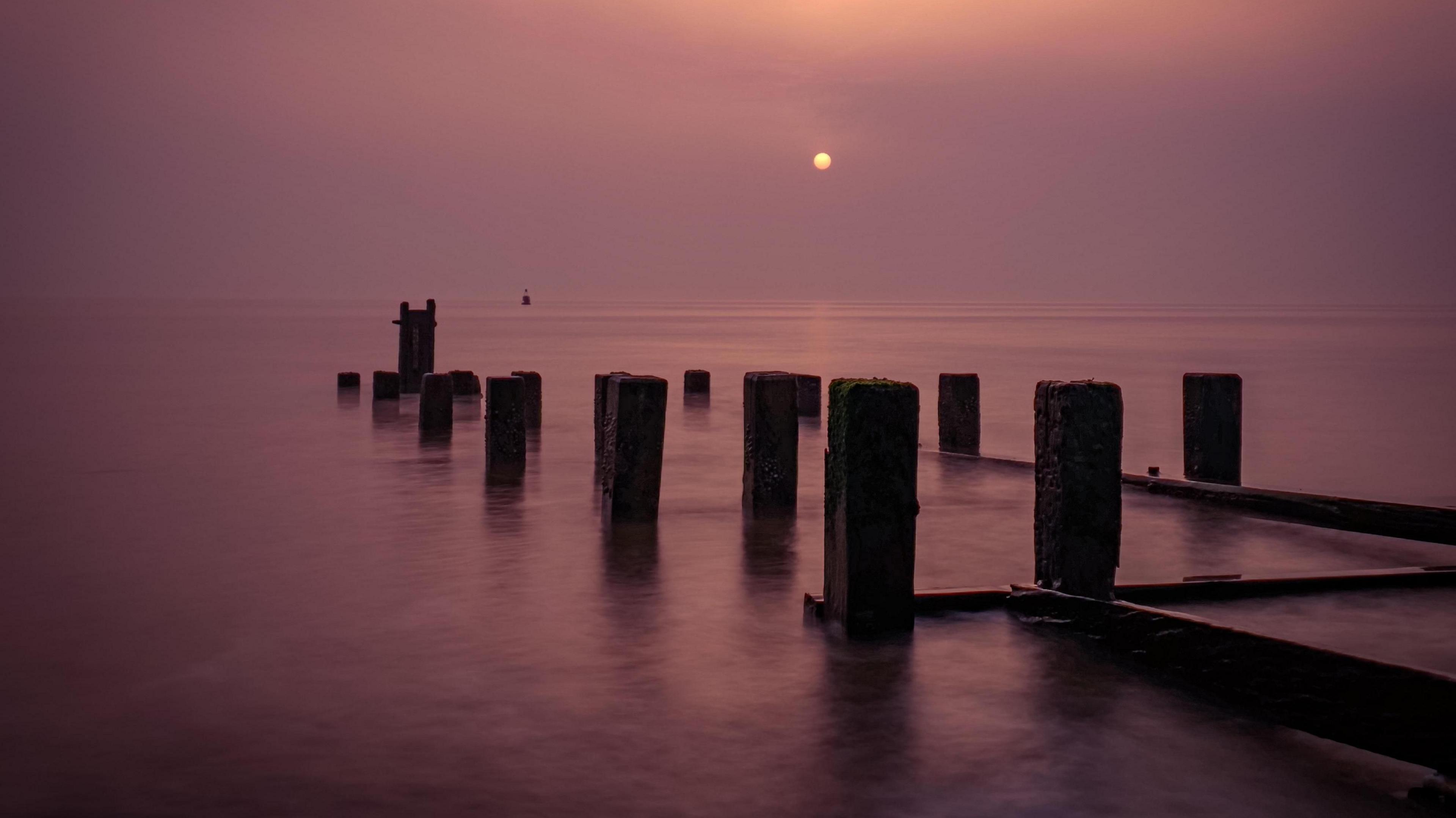 Wooden groynes jut out into a pink sea reflecting the pink dust-laden sky 