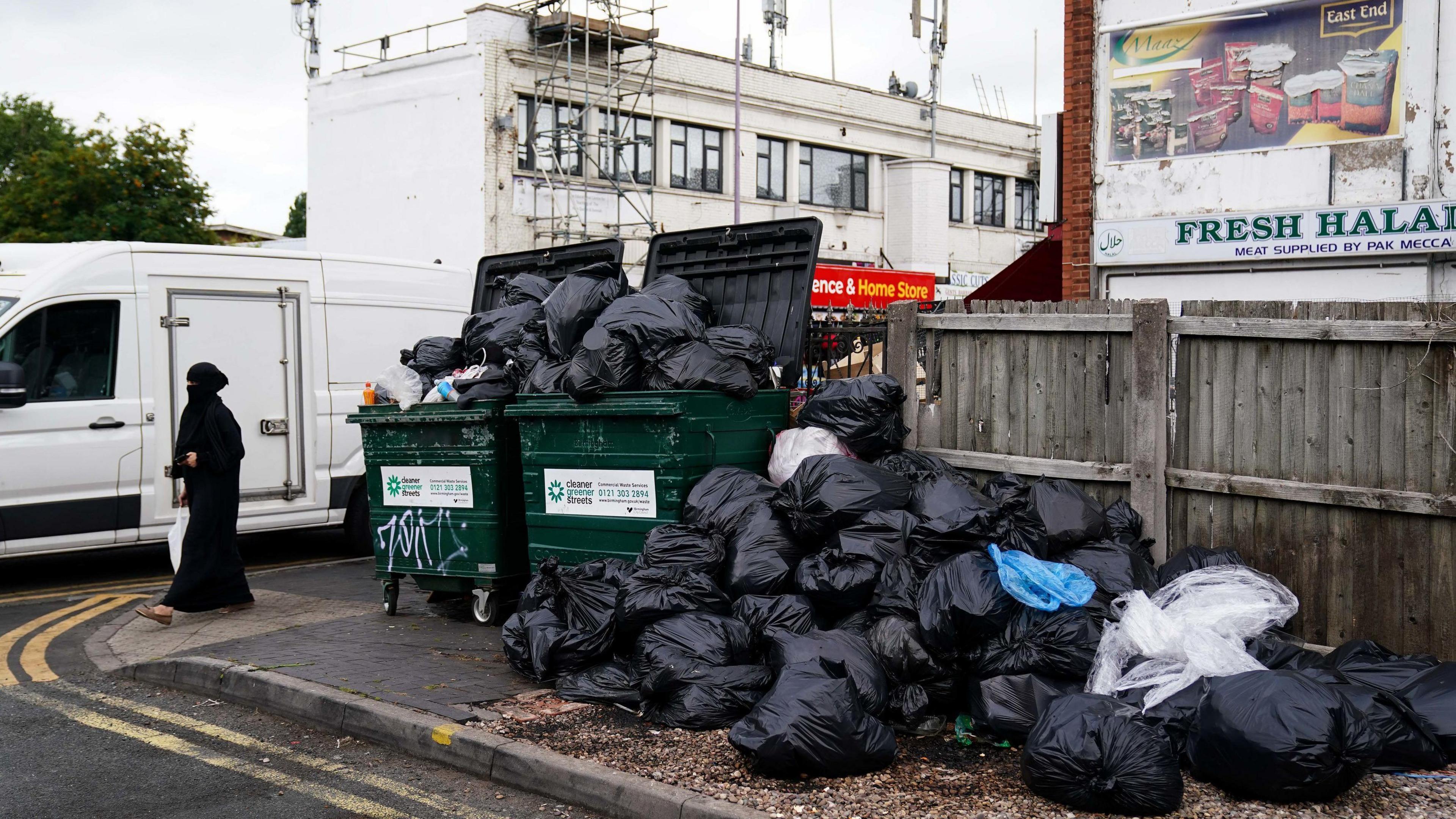 A woman dressed in a black face covering walks past over flowing green bins with black back piled up high.