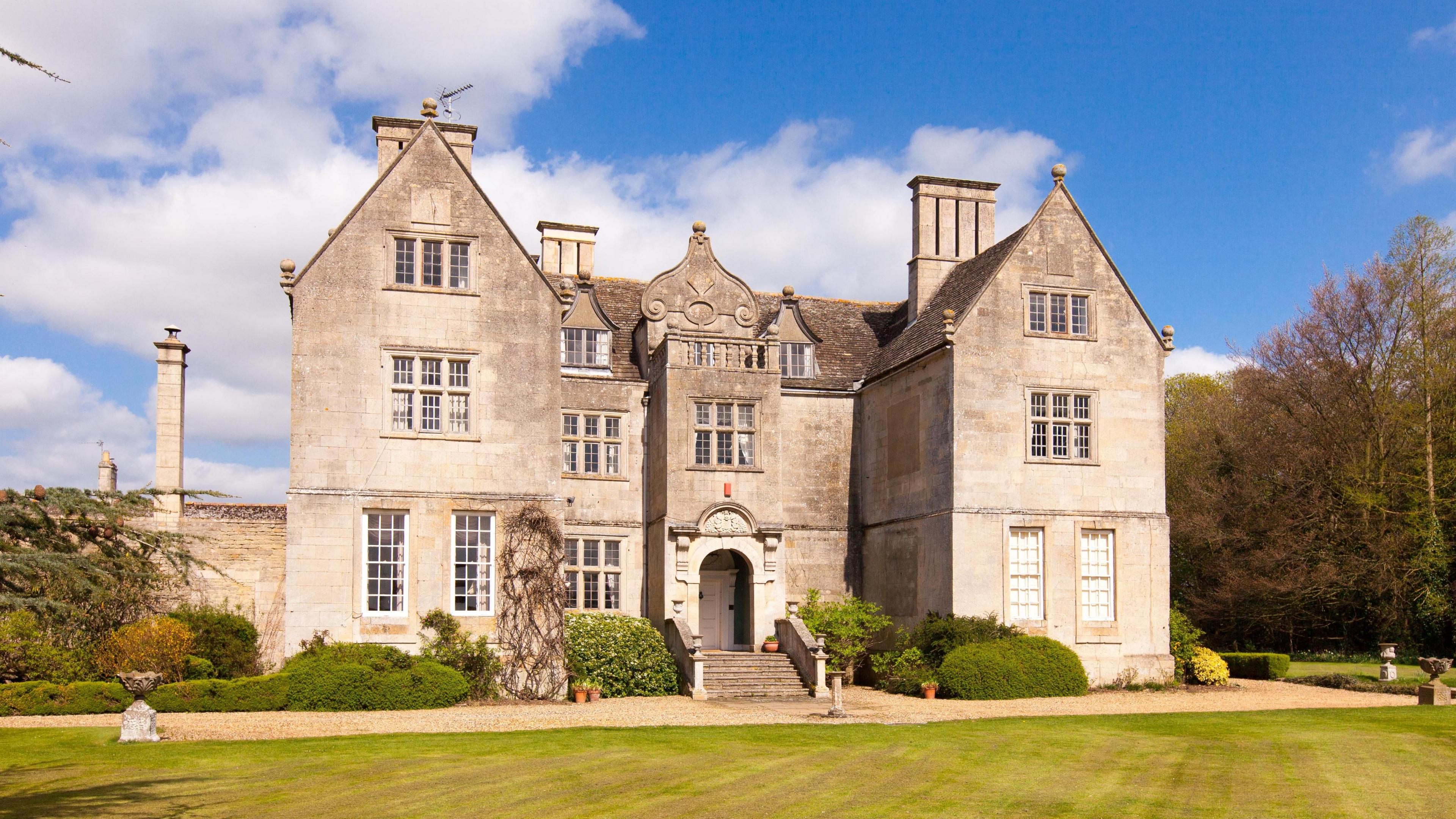 A grand, faded brown brick building stands in front of a mowed green lawn. There is an arched doorway and steps leading up to it, trimmed hedges and a tree to the right. The sky is blue with white clouds, the weather sunny.