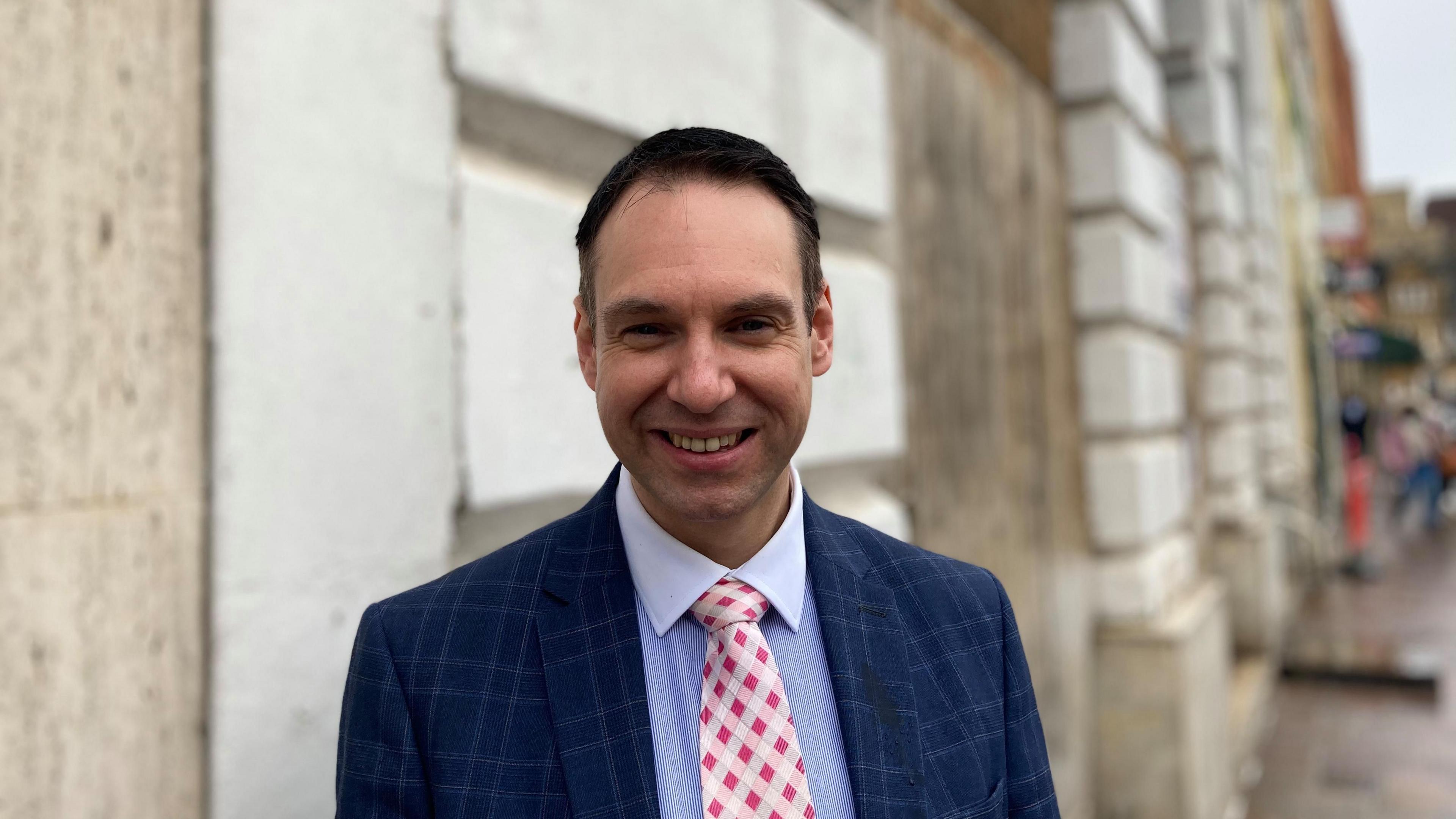 Councillor Dan Lister in a striped royal blue shirt, a pink checked tie and a navy checked jacket. He has brown haired, styled in  a short back and sides do and is smiling at the camera. The background is out of focus but his is stood in the town centre in front of a beige building