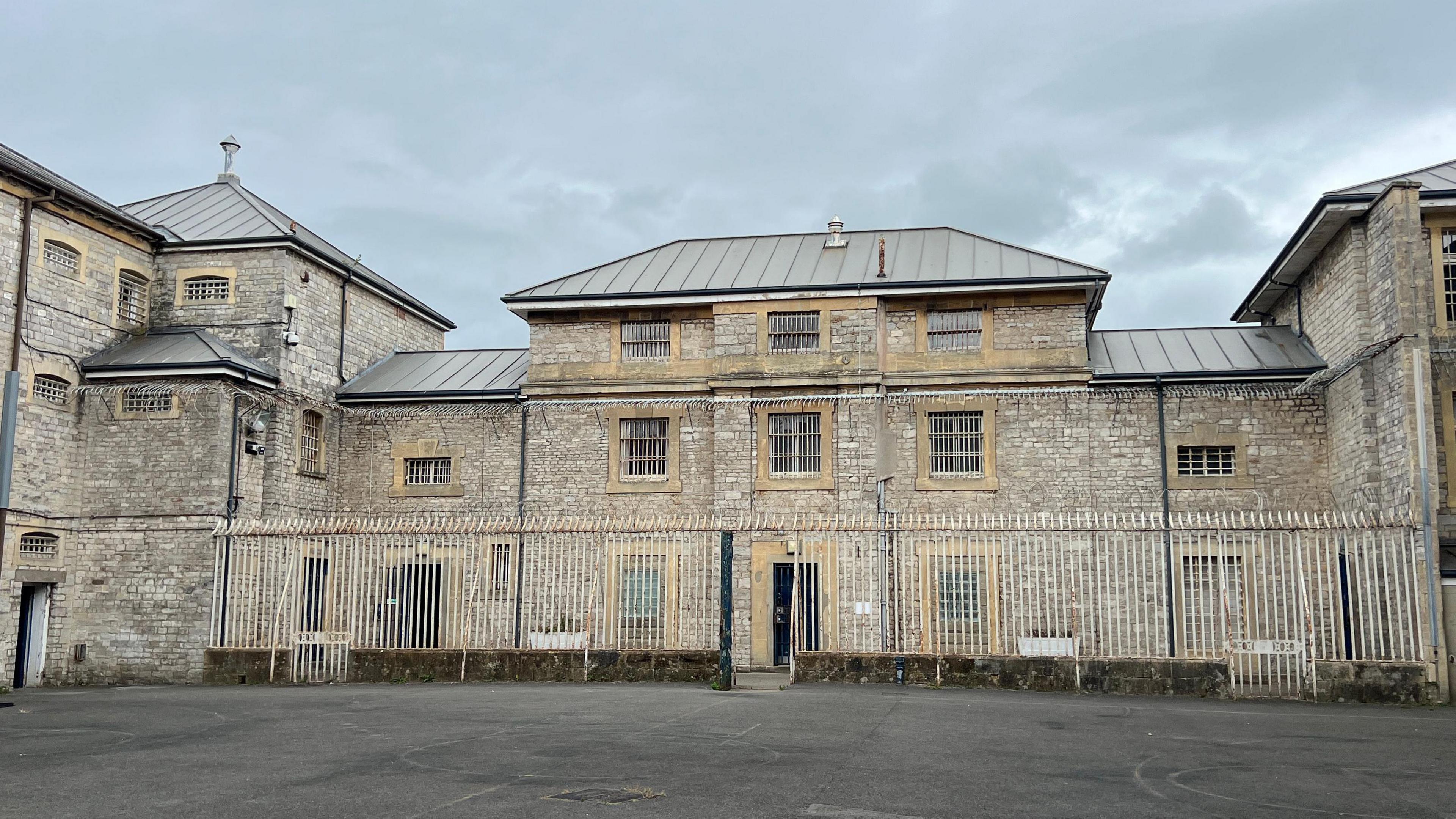 A large grey stone prison building with white gates sits under a gloomy sky.