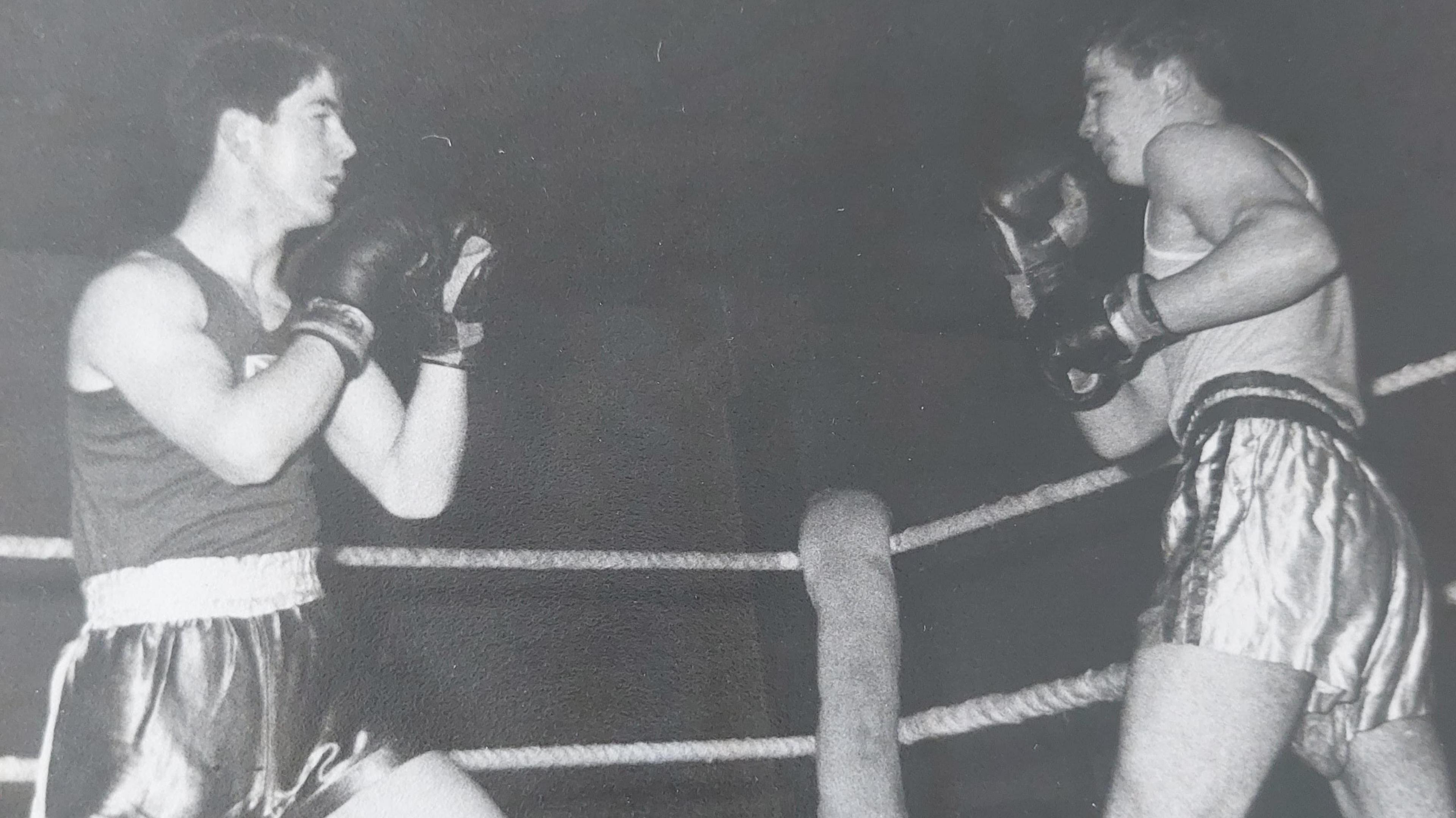 A vintage black and white photo of a young Frank Gilfeather wearing boxing shorts and a tank top with the Scotland flag in the centre, in a boxing match against a taller man