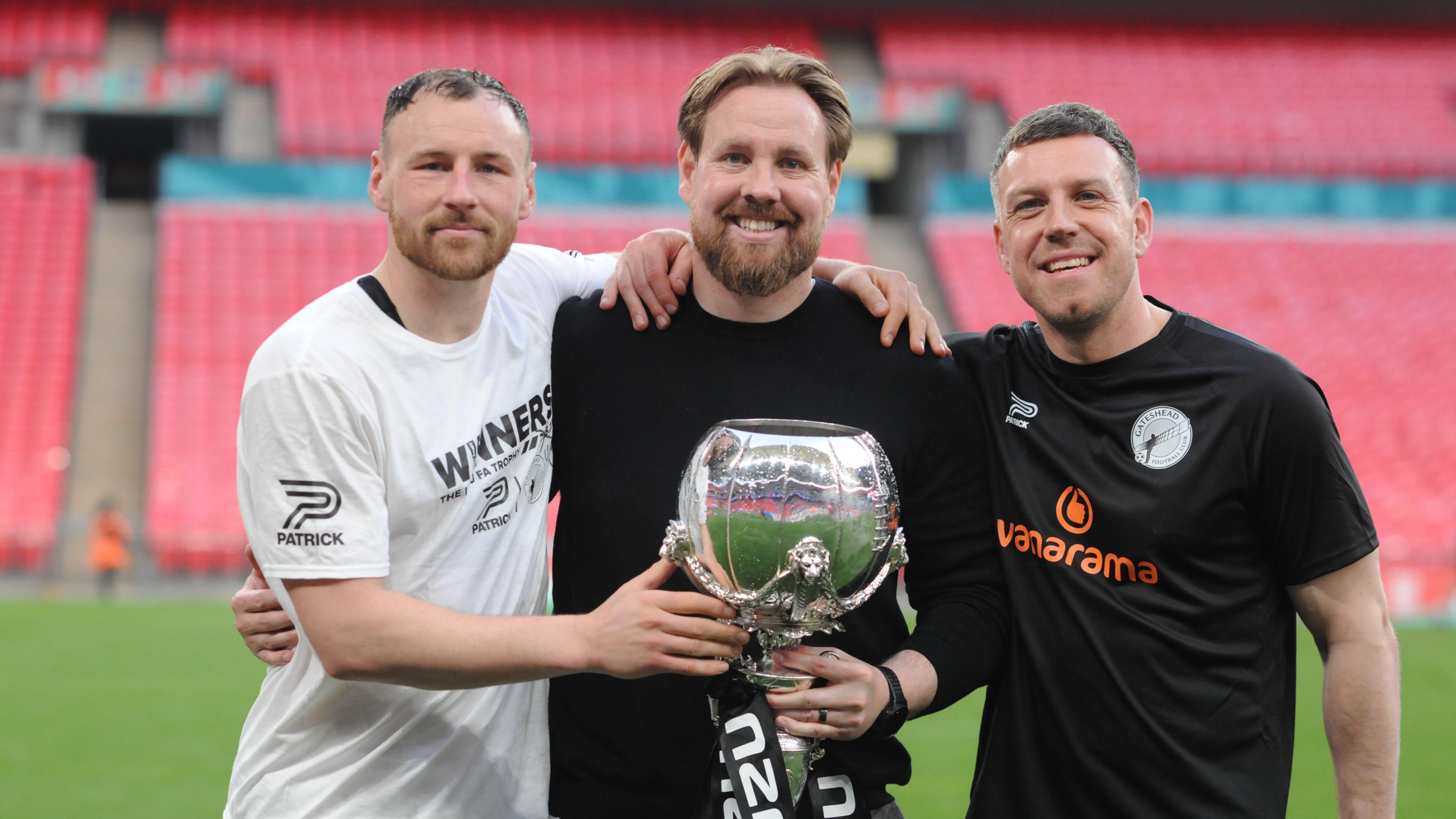 Rob Elliot holding the FA Trophy at Wembley in 2024 flanked by a player on his left and Carl Magnay (right)