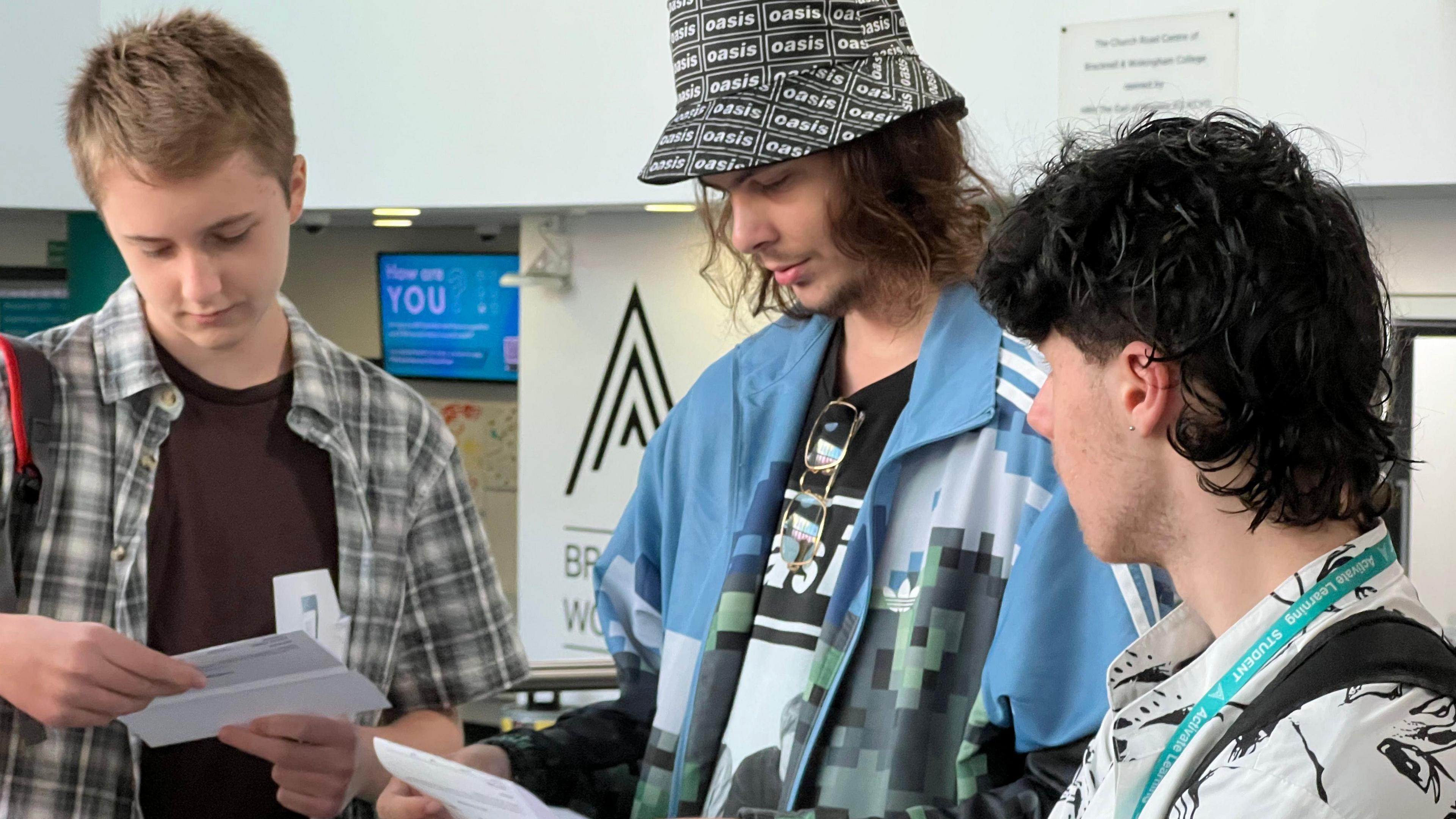 Three young men look at pieces of paper with their results on. The man to the left has short blonde hair and wears a brown t shirt with a brown and grey check short sleeved shirt over the top and a single rucksack strap hooked over his shoulder. The student in the centre in a man with mid length brown hair and a short beard. He wears a black and white bucket hat which says "Oasis" on it and wears a short sleeved Adidas shirt with an Oasis band t shirt underneath. The student to the right has a black mullet and wears a white and black printed shirt.