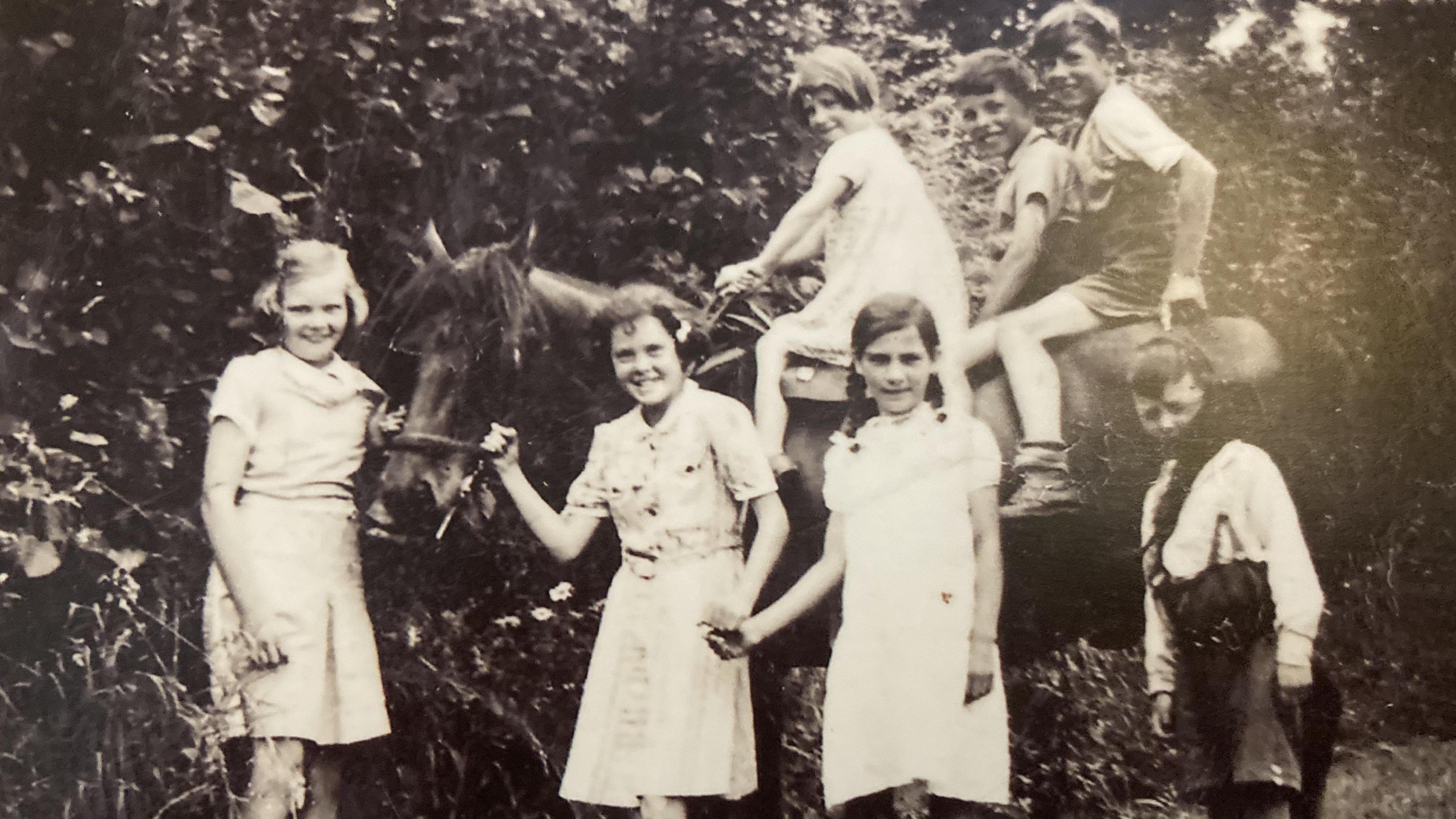 This is a black and white photo. It shows children standing in front of a horse. Three more children are sitting on the horse. The photo was taken in 1941.