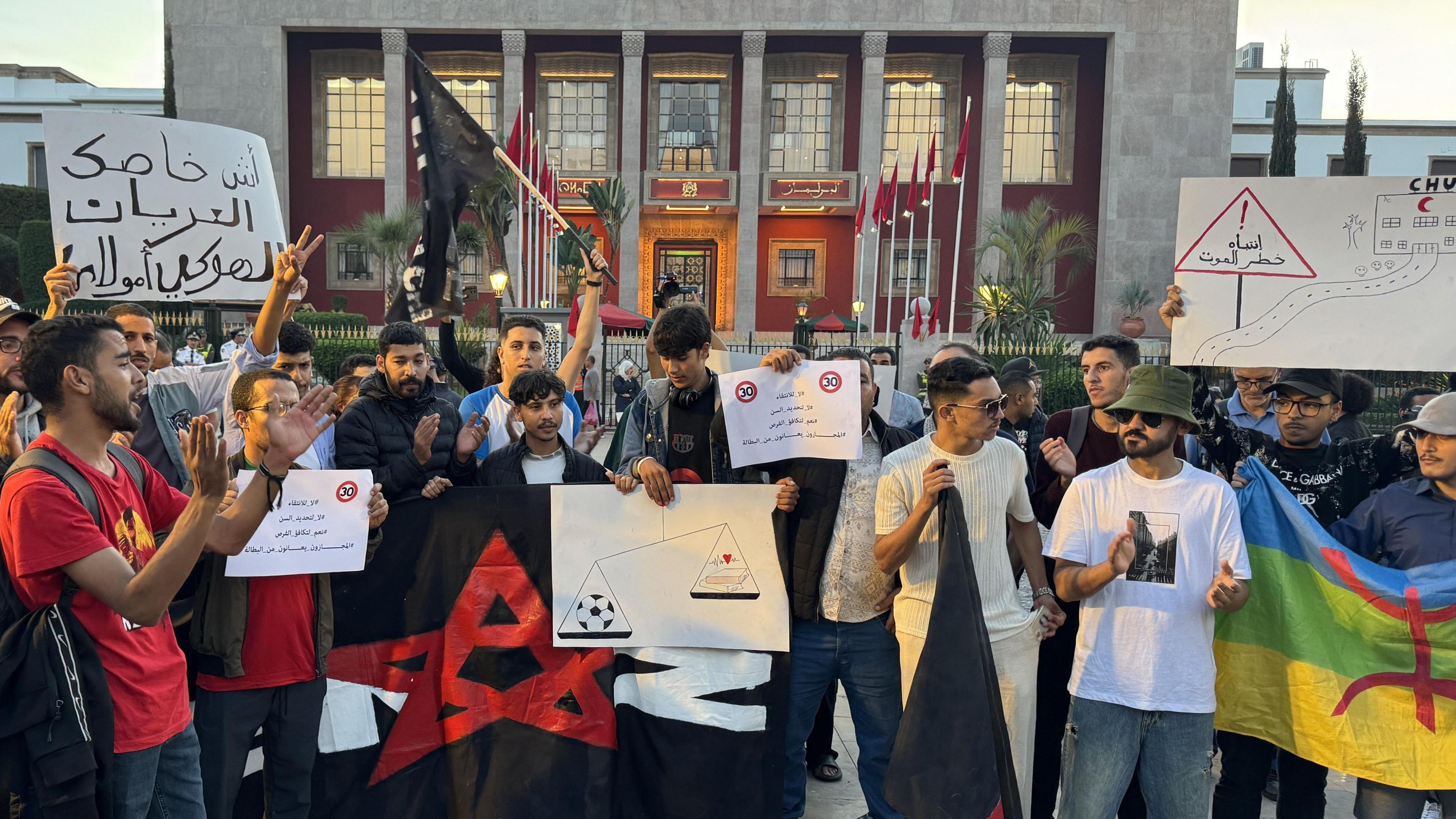 Gen Z protestors in Morocco stand behind banners and hold up placards in front of an administrative building in Rabat