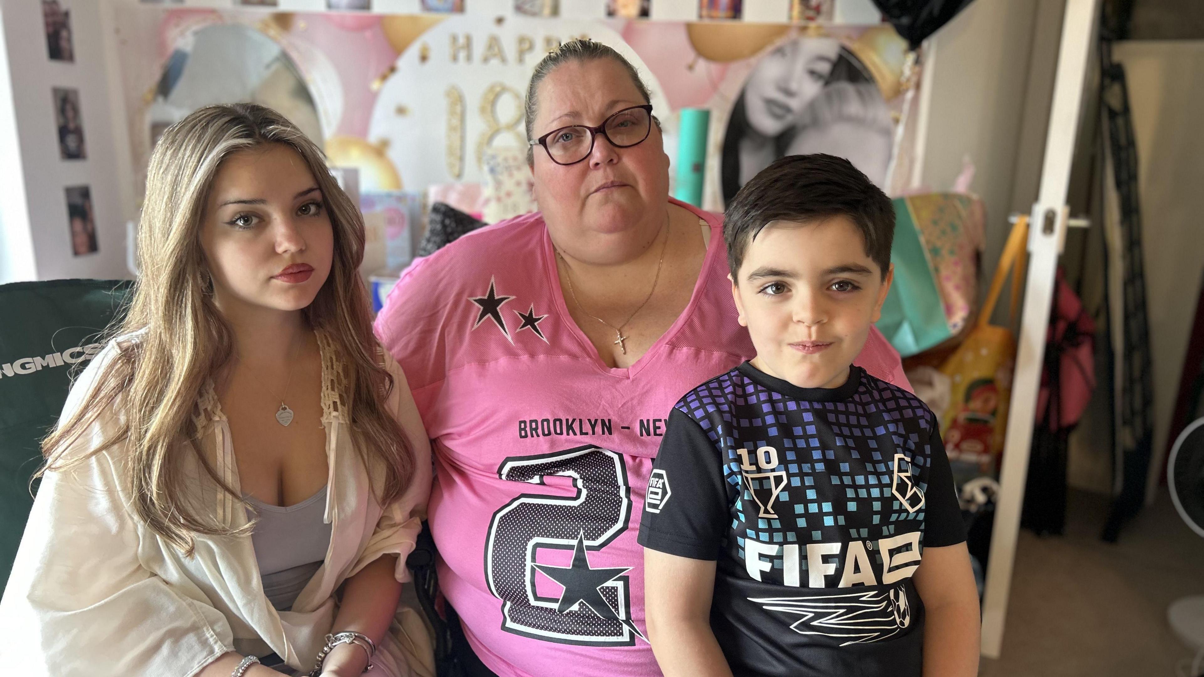 A mother wearing a pink printed t-shirt sits between her two children, an 18 year old wearing a white shirt and a 6 year old wearing a black and blue FIFA top