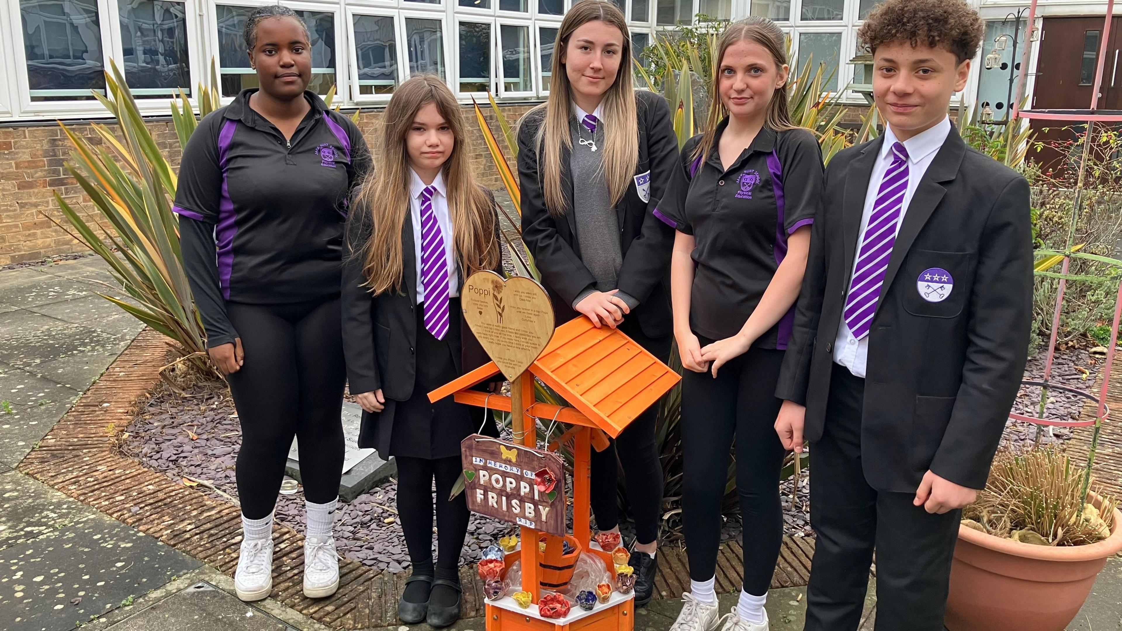 Poppi's friends, four girls in black and white school uniform and a boy, also in uniform, standing in front of an orange wooden memorial with clay poppies. The memorial is in a garden. It is surrounded by school buildings.