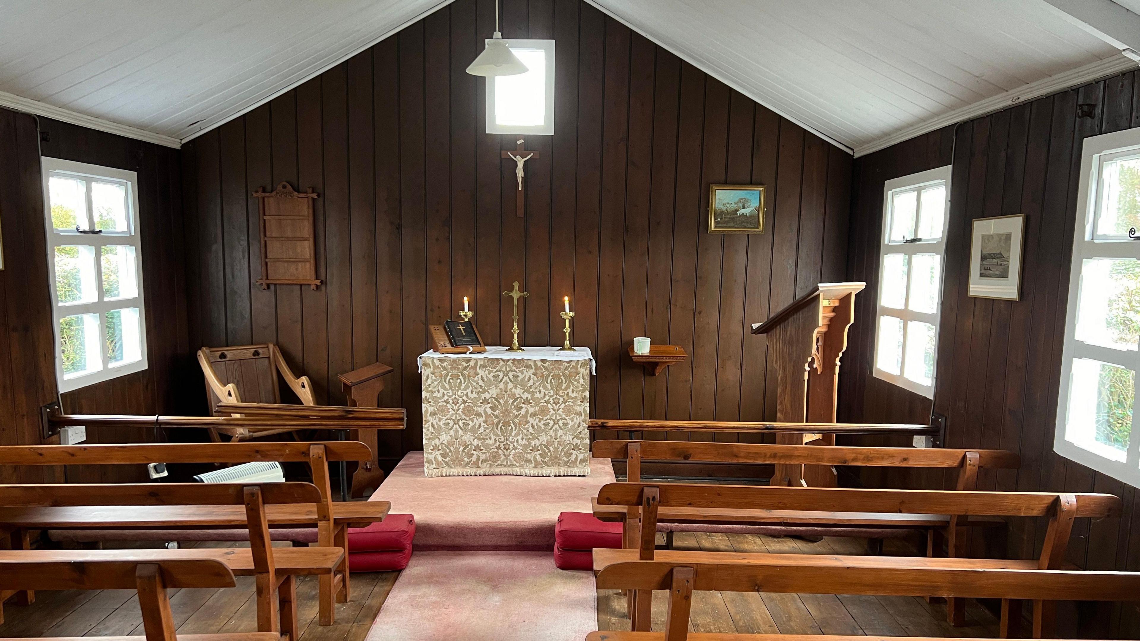 The inside of the church. It has wooden walls and there are wooden pews. The altar has candles on it that are lit.