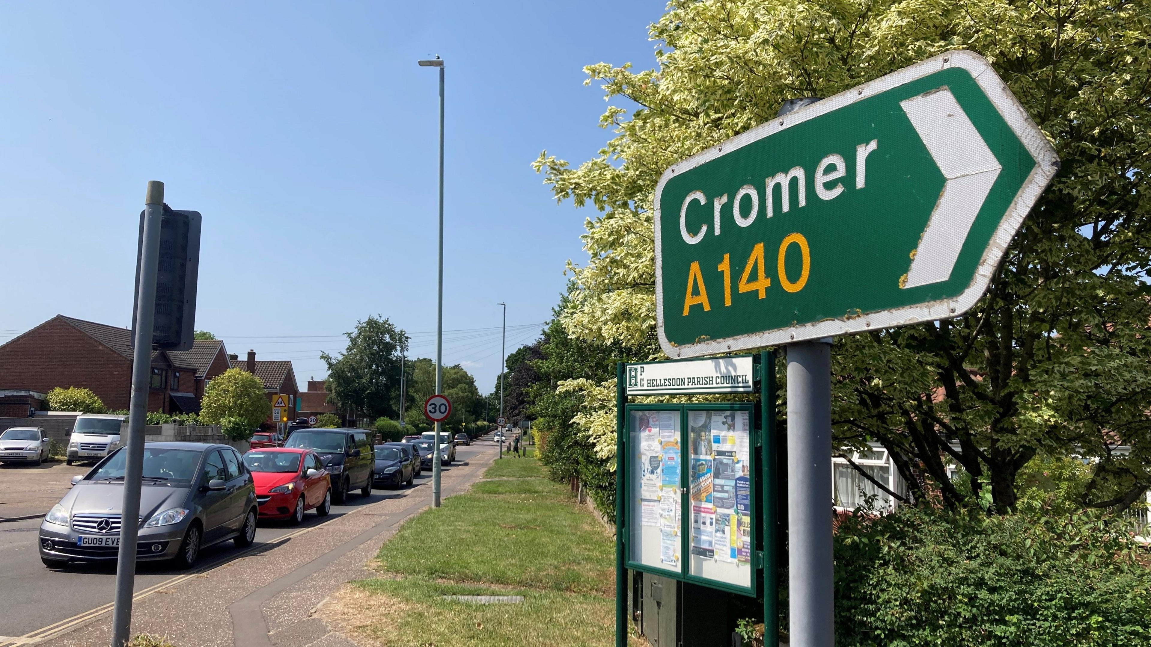 A green signpost with writing on it saying A140 to Cromer. Traffic is stationary in the background while it waits at traffic lights.