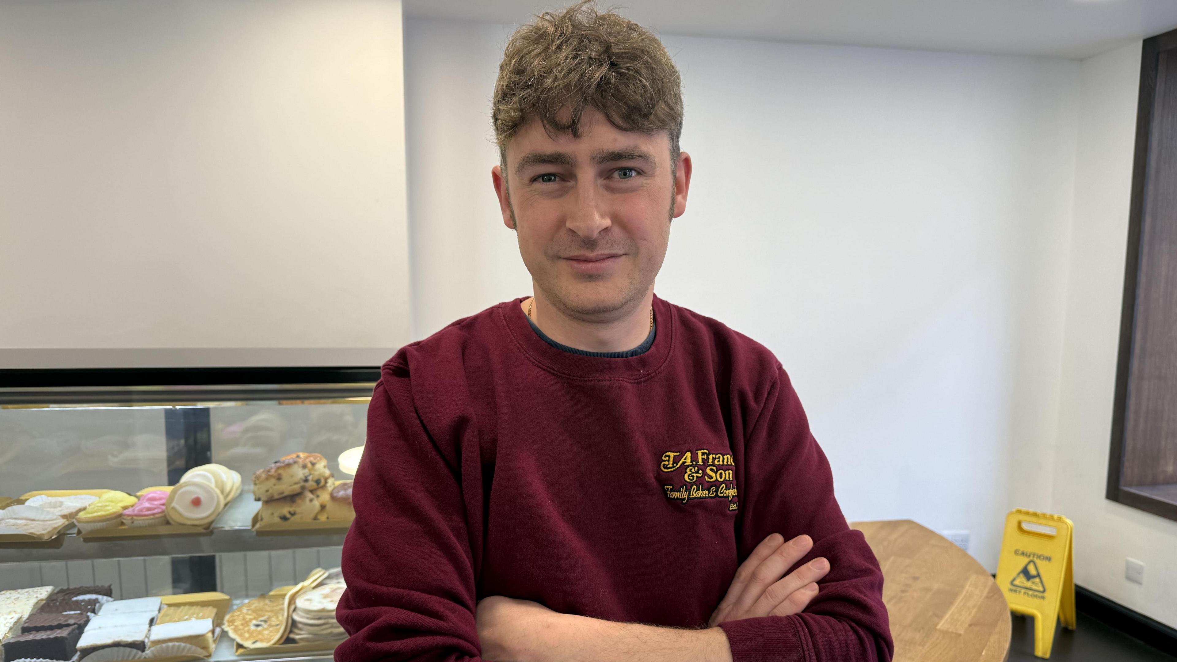A man stands with his arms crossed, in front of a bakery counter filled with baked goods. He has short brown hair and is wearing a red jumper which says "T.A. Francis & Son."