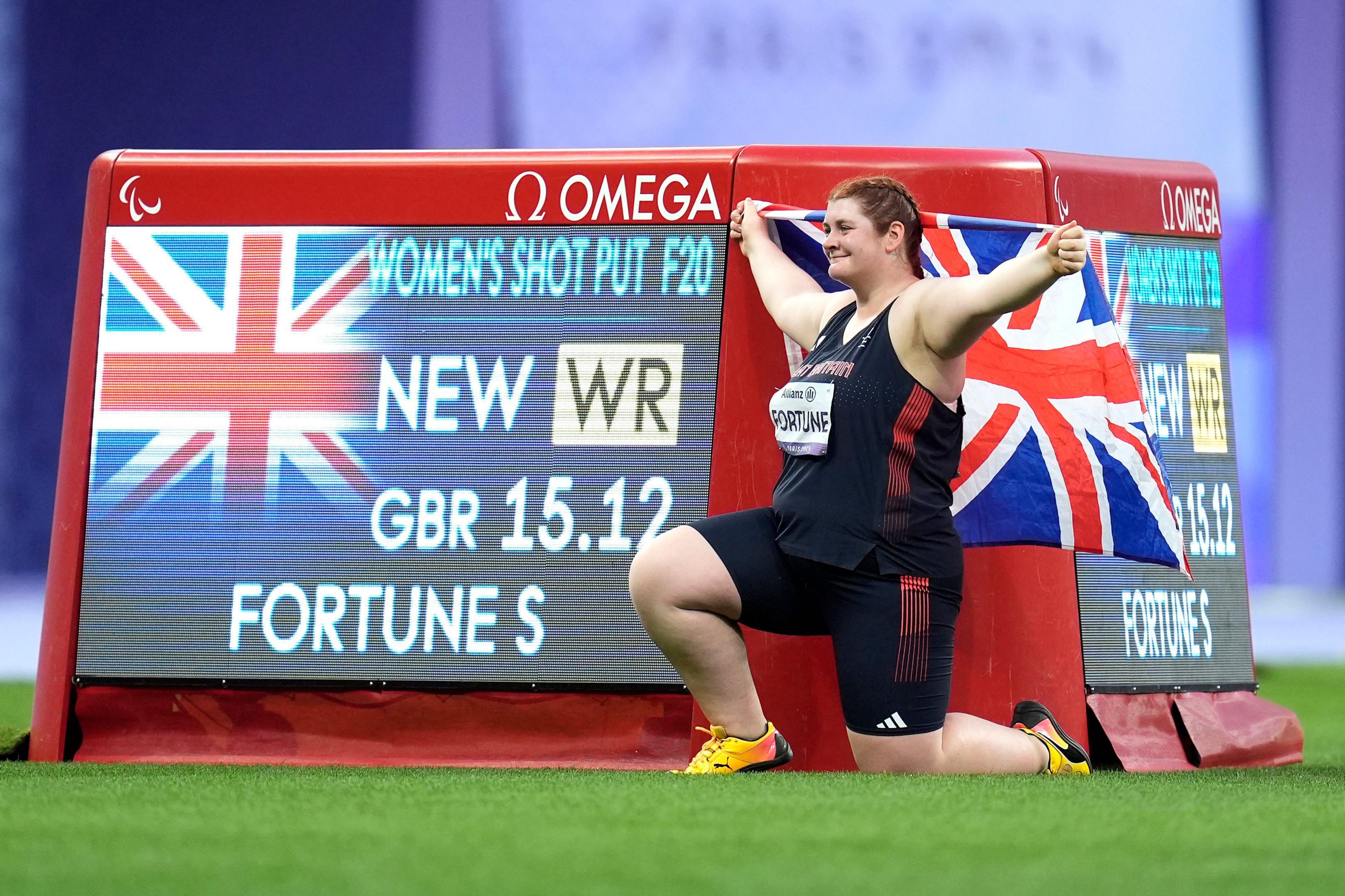 Sabrina fortune kneels next to her world record achievement displayed on a screen while holding up the flag of Great Britain looking proud