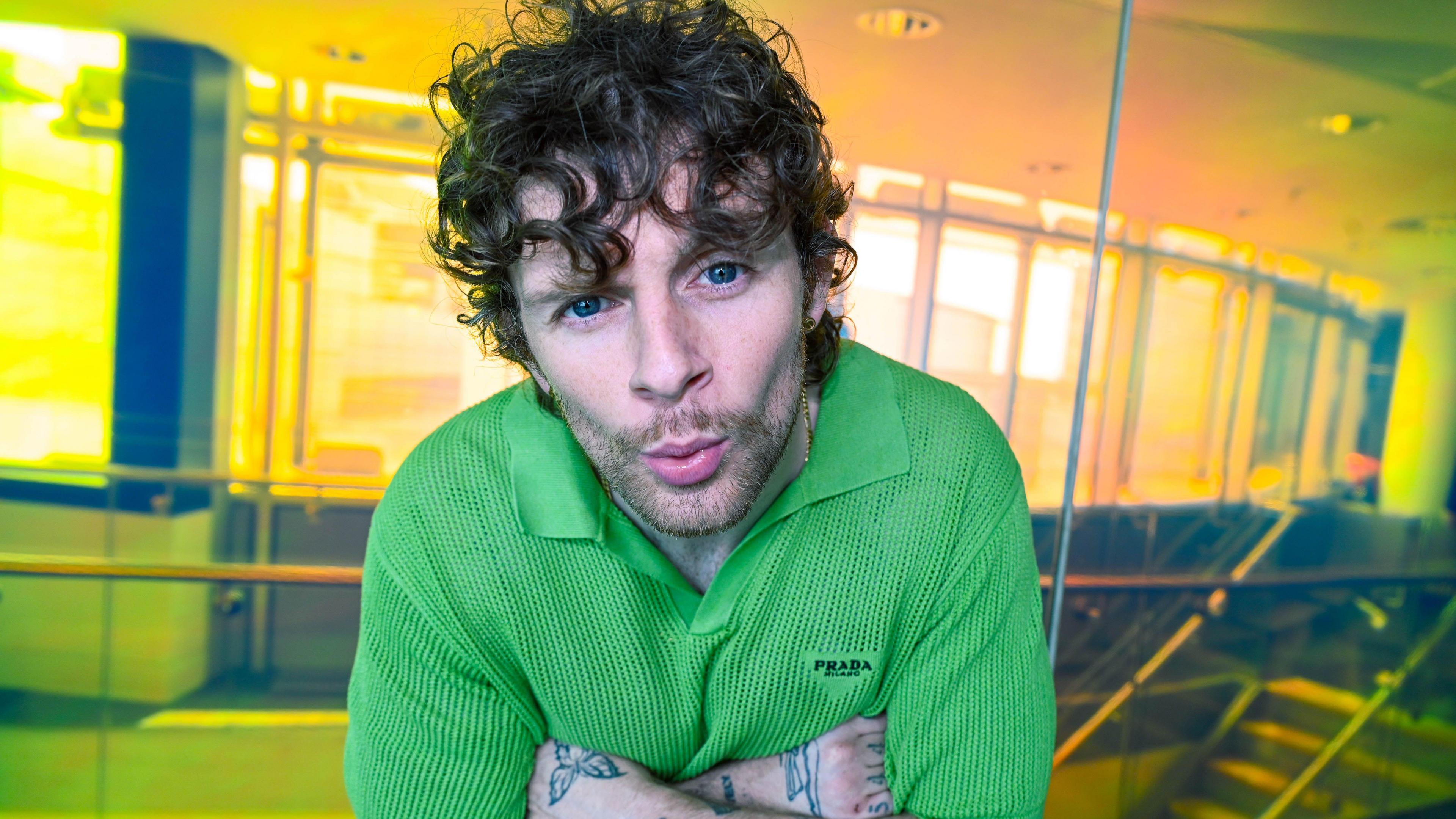A man with curly hair and visible tattoos on his arms is wearing a green collared Prada shirt. They are indoors in a space with glass walls and warm yellowy/orange lighting. The man is leaning slightly forward, facing the camera and is pouting his lips.