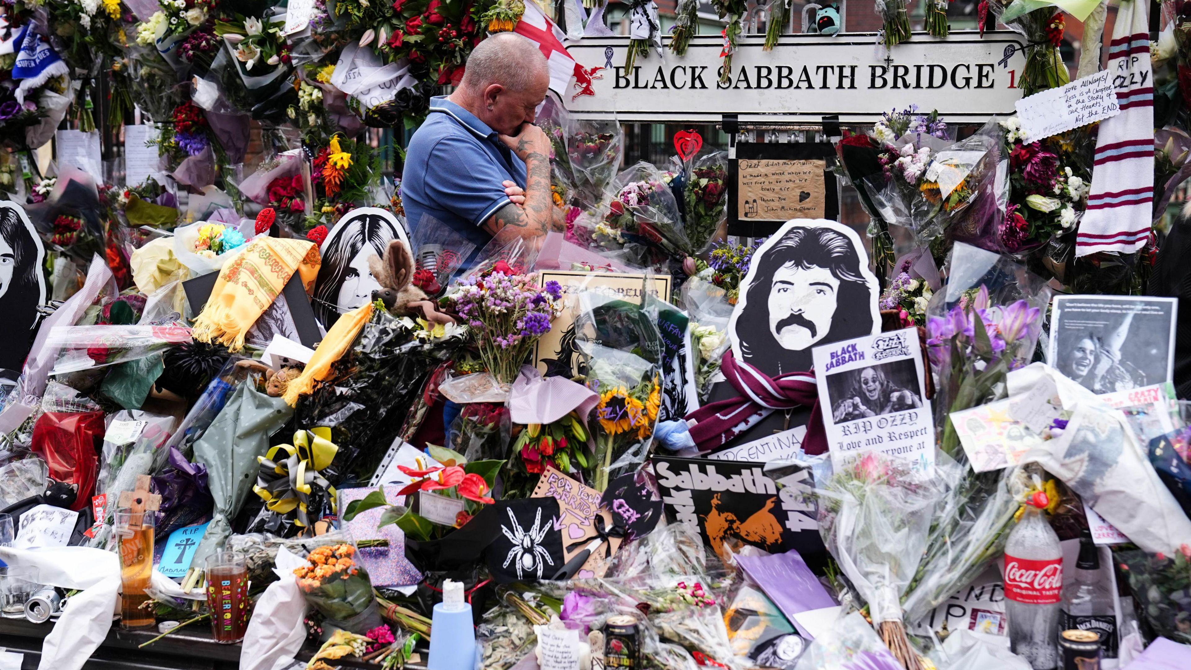 A man stood surrounded by flowers and notes and a sign saying Black Sabbath Bench