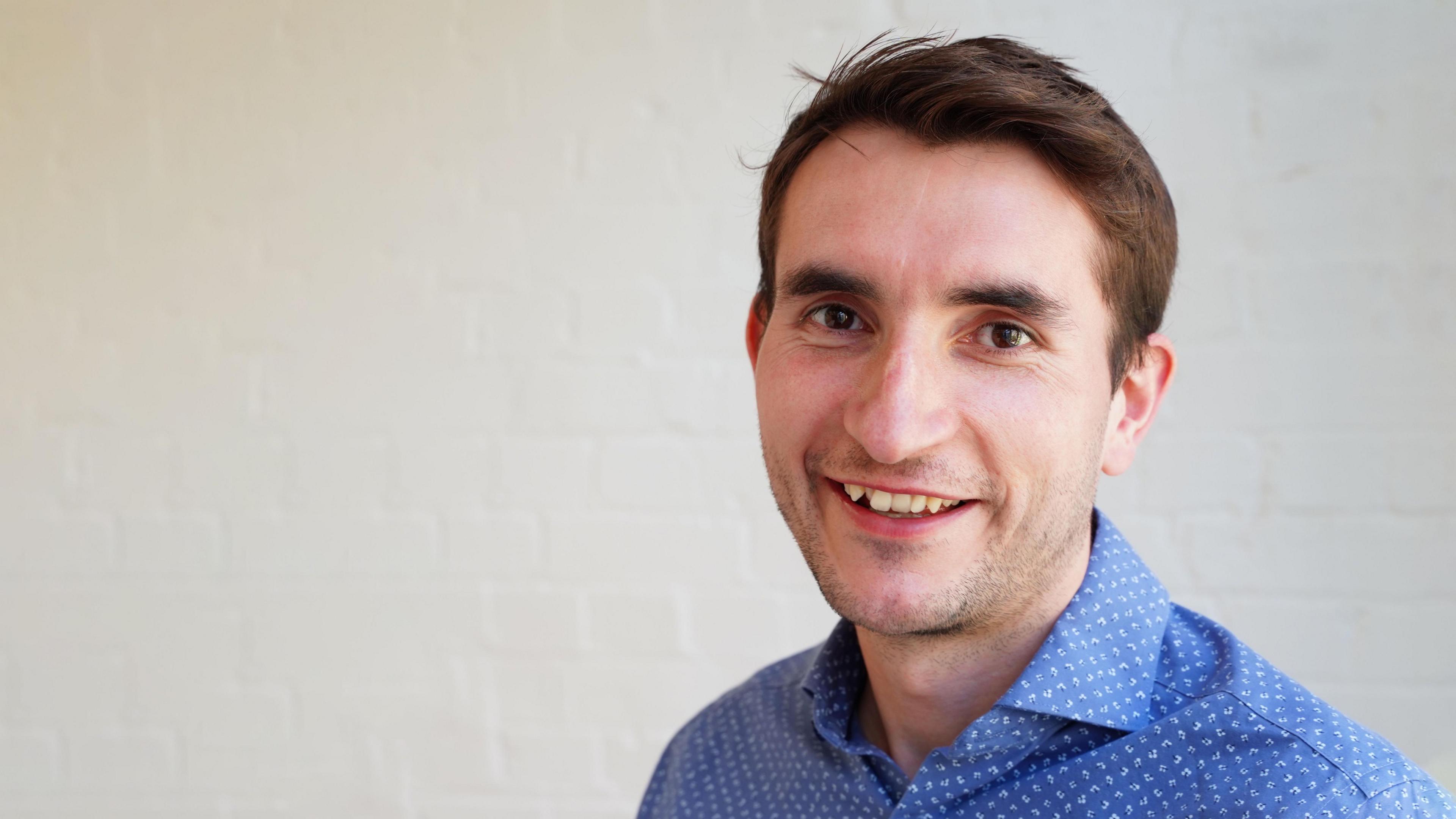 An off-centre Paul Swinney is in front of a white painted brick background, wearing a white and blue shirt, smiling at the camera.