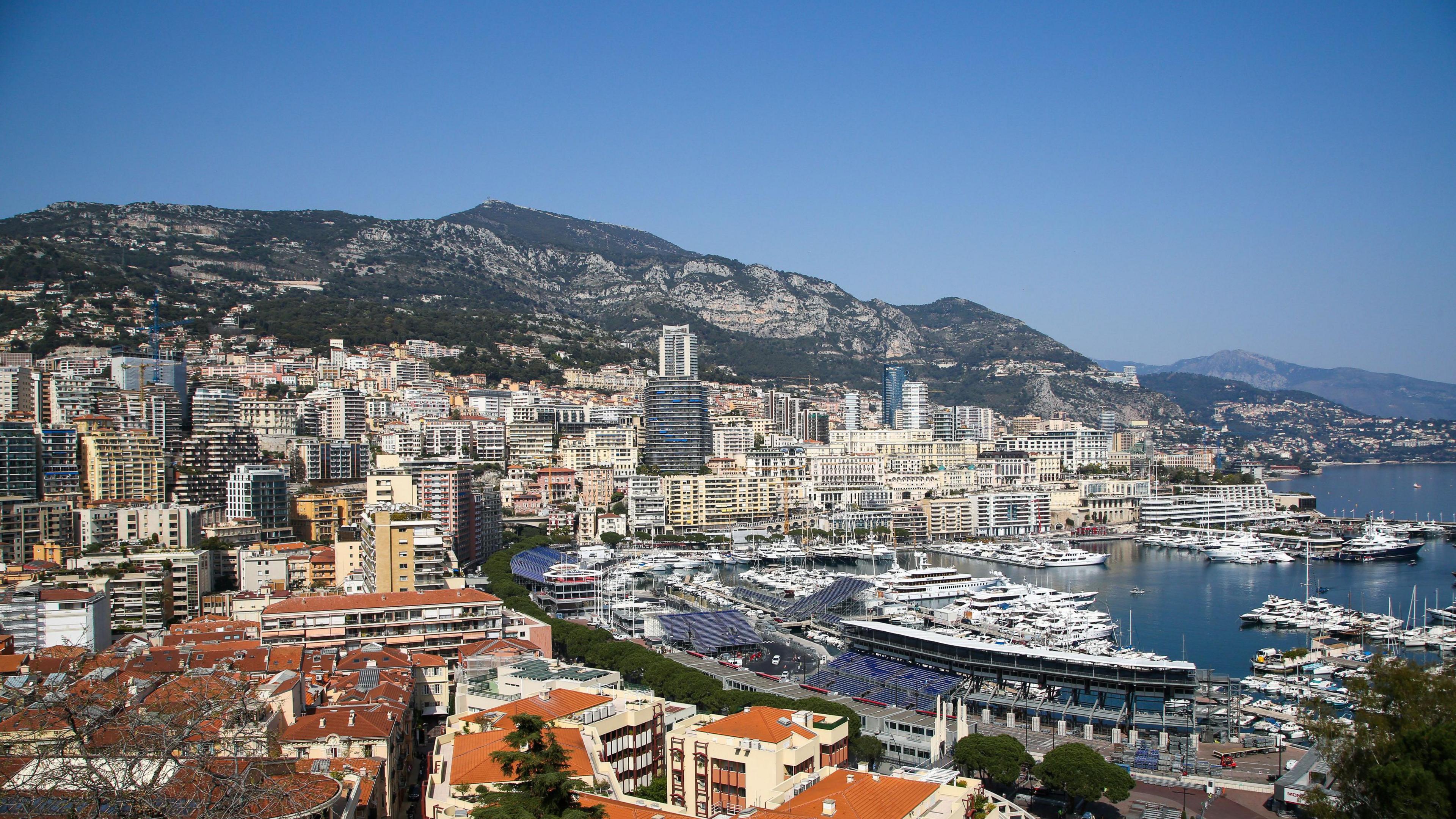 Aerial view of Port Hercules, filled with yachts, and the tower blocks of Monte Carlo in Monaco, with mountains rising behind them and Cap Martin visible in the distance