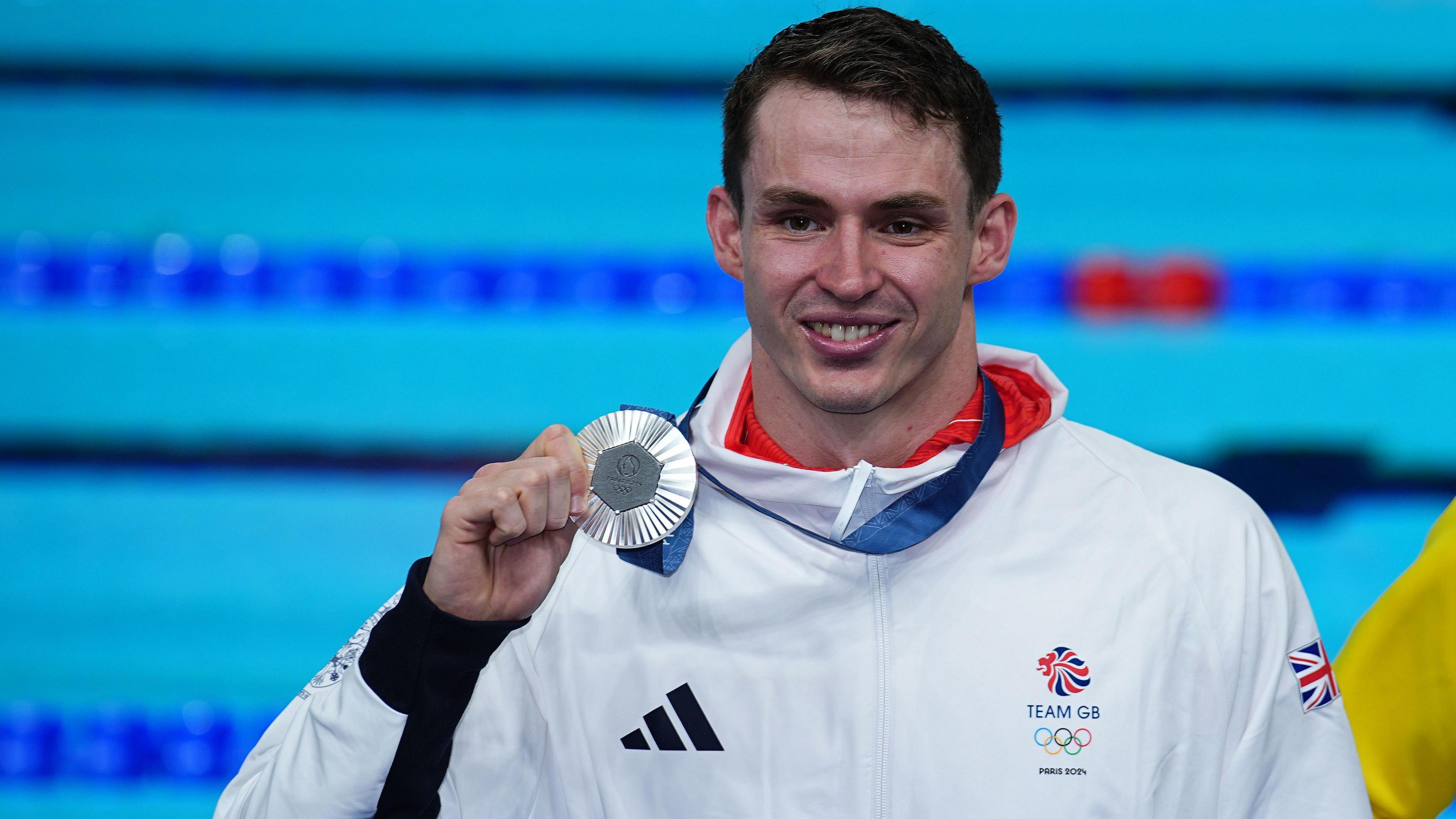 Swimmer Ben Proud holds his Olympic silver medal in his right hand while he stands next to a swimming pool wearing his white Team GB jacket.