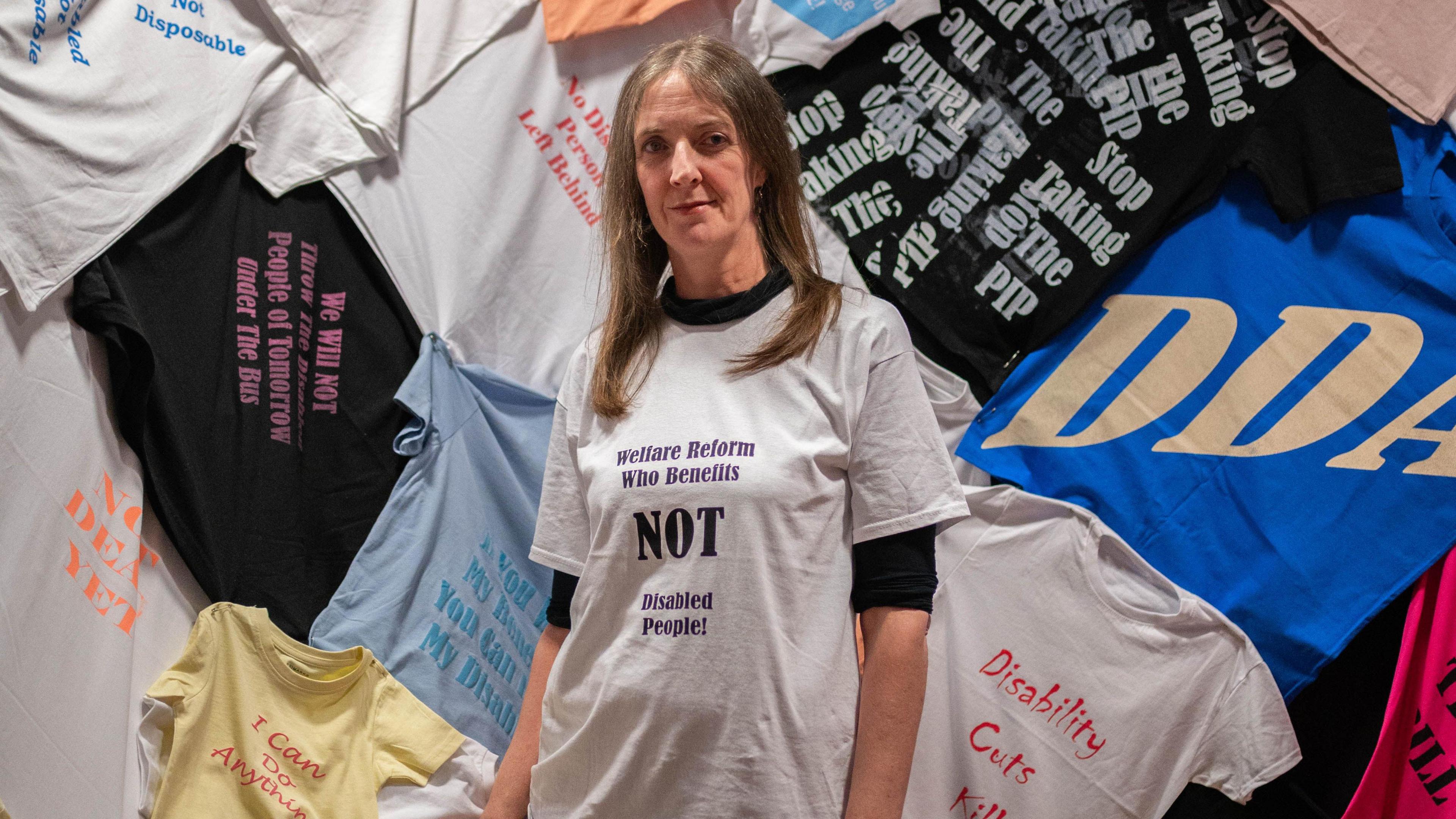 Fae wearing a white T-shirt and a black full-sleve inner shirt standing in front of the T-shirt display, smiling for camera.