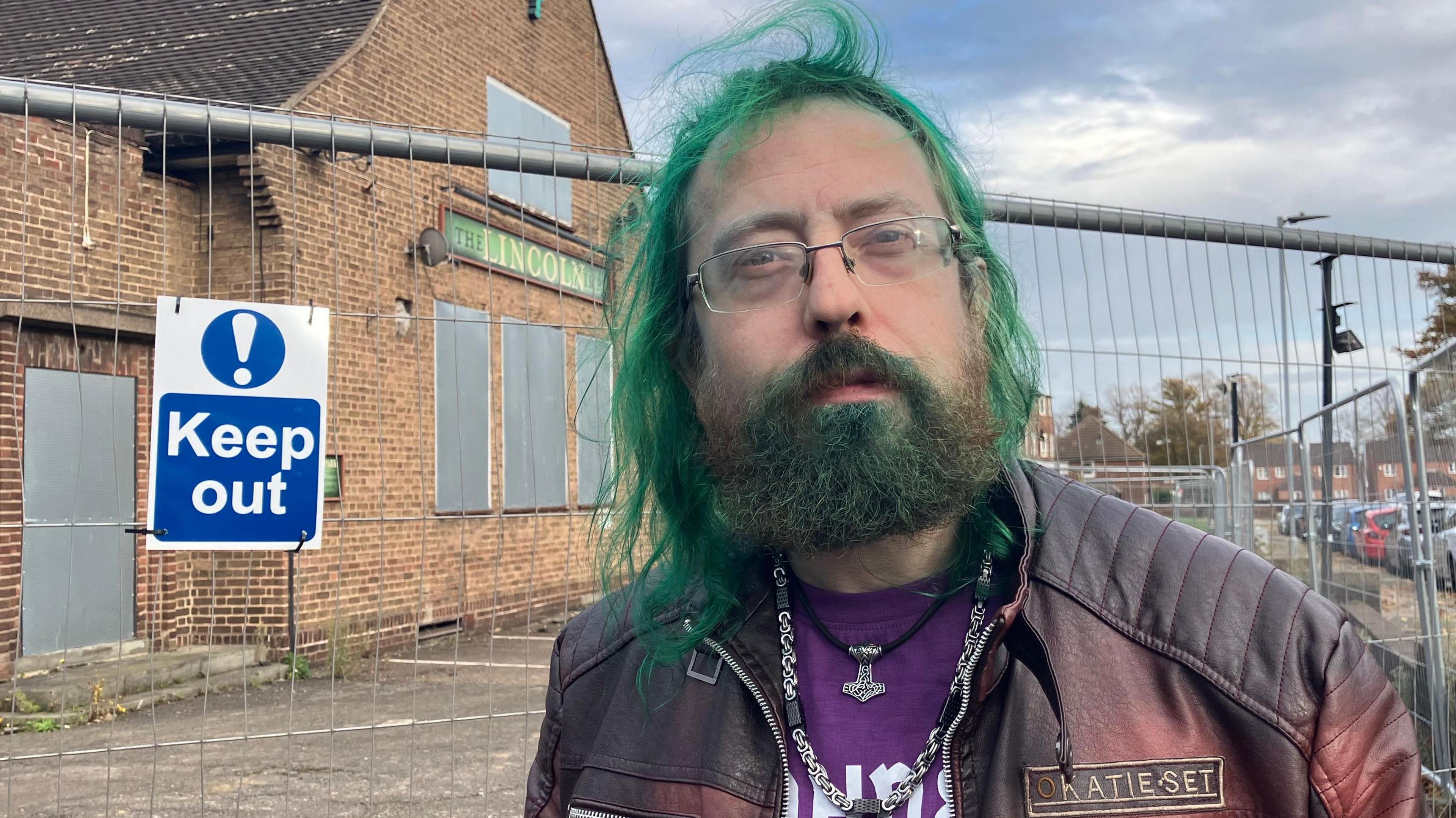 Malachi Piatt has long green hair, a beard and glasses. He has a brown jacket and a purple top. He stands in front of a derelict building with a sign that says "keep out".