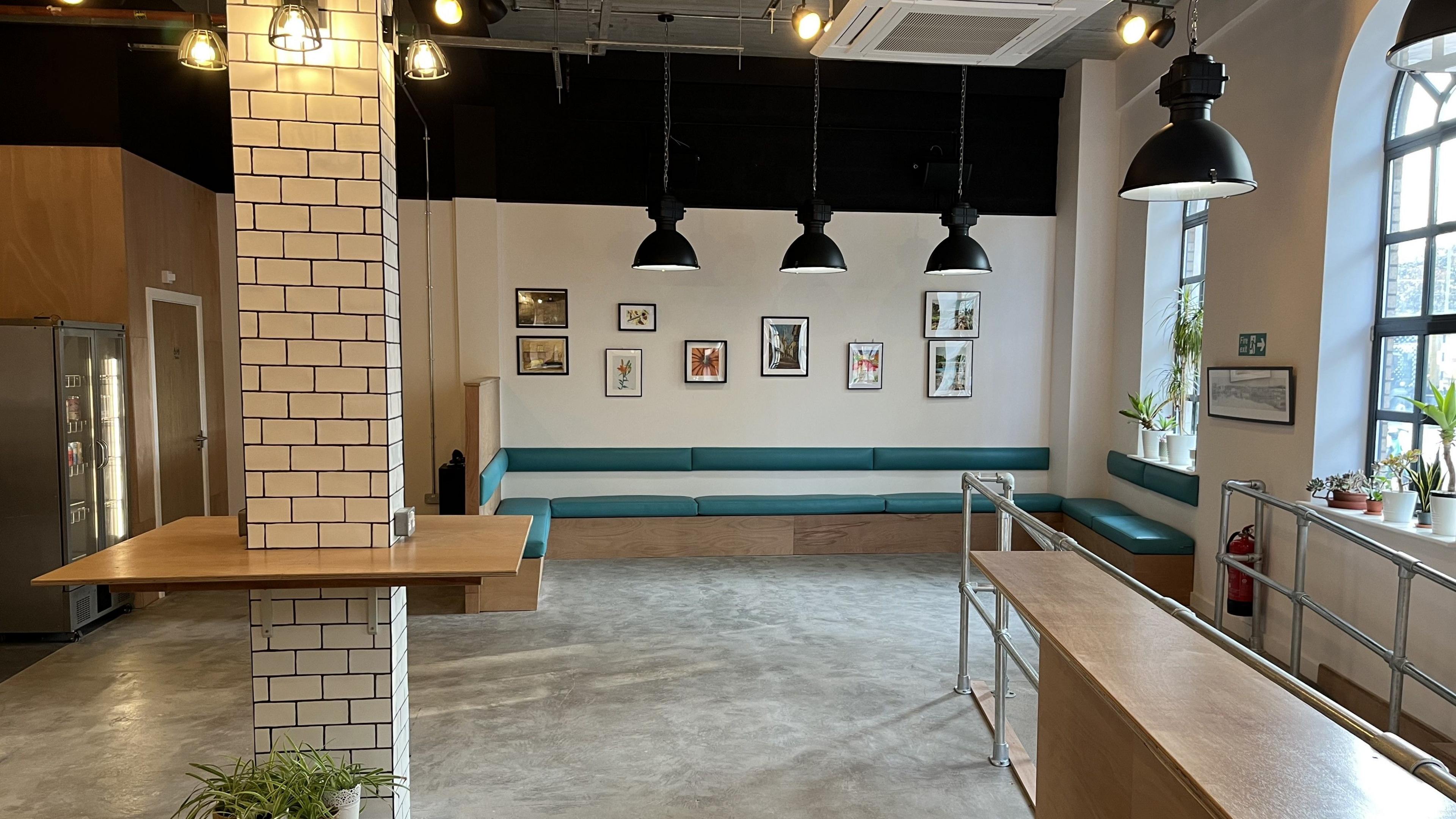 The interior of the newly refurbished Key Cafe. It is a modern building with a slightly industrial feel. The ceilings are high with hanging overhead lamps, and there are large arched windows on the right letting plenty of light in. There is a sofa on the far wall with framed pictures hanging above it. There is a tiled column in the middle of the room with a wooden table around it. On the right there is a ramp leading outside with metal barriers either side. 