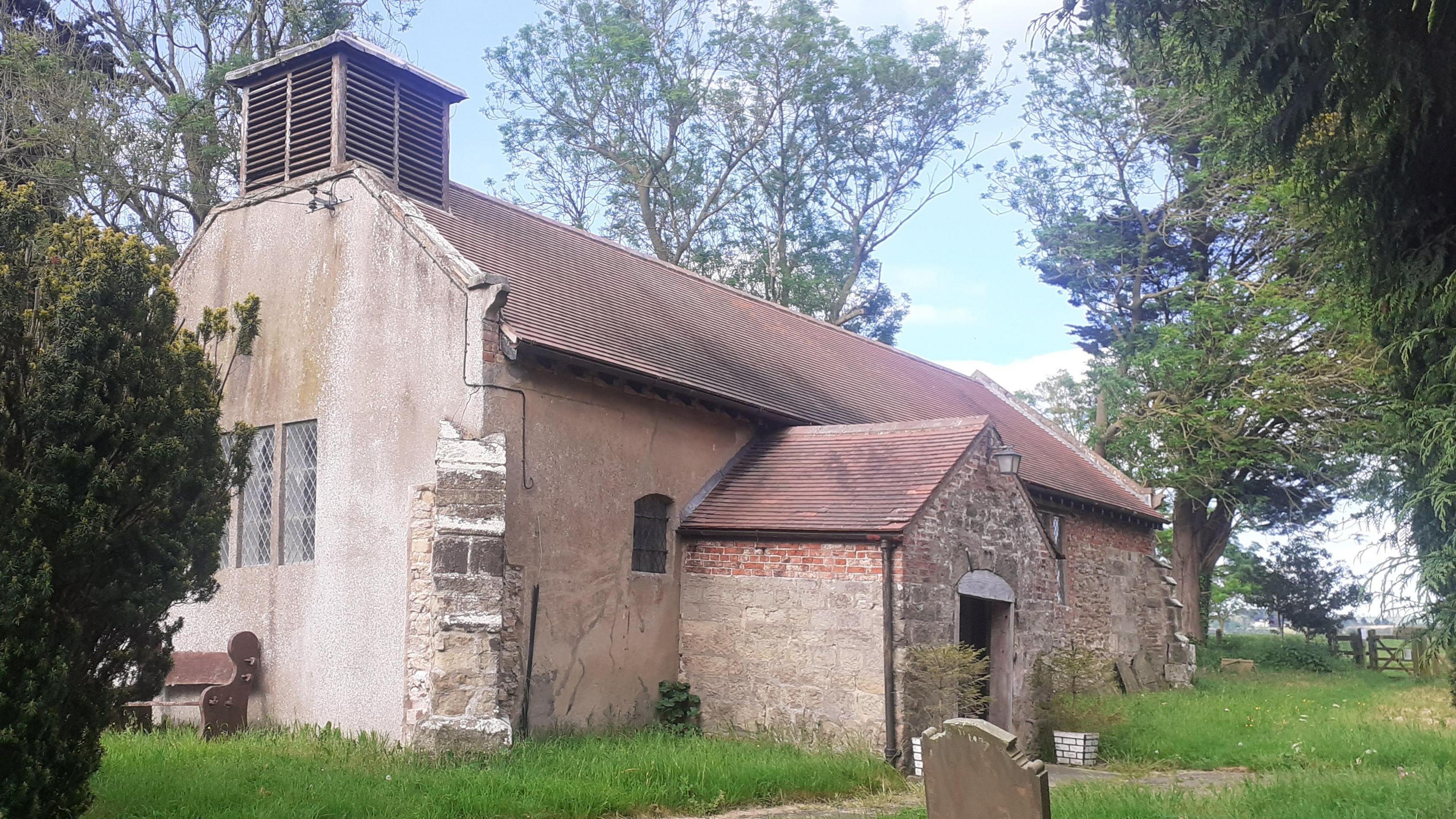 The churchyard at Yapham. The main church building is in the centre of the image with a large church tower in the background. A graveyard is visible in the foreground with large shrubs and iron railings surrounding the site.

