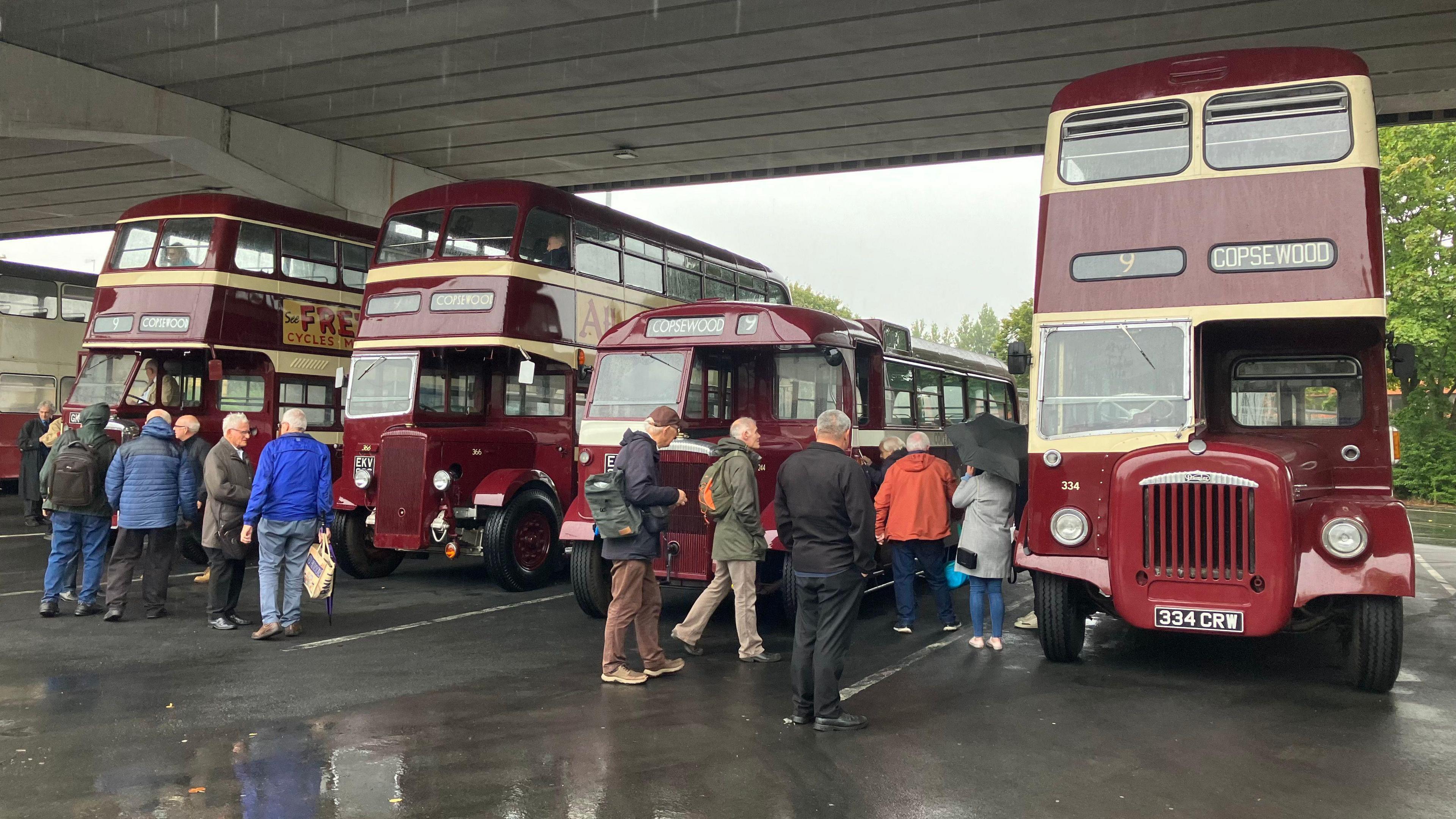 Groups of people stand outside four vintage red buses, three of which are double-deckers and one of which is a single-decker. They stand under a bridge, with water gleaming on the tarmac.