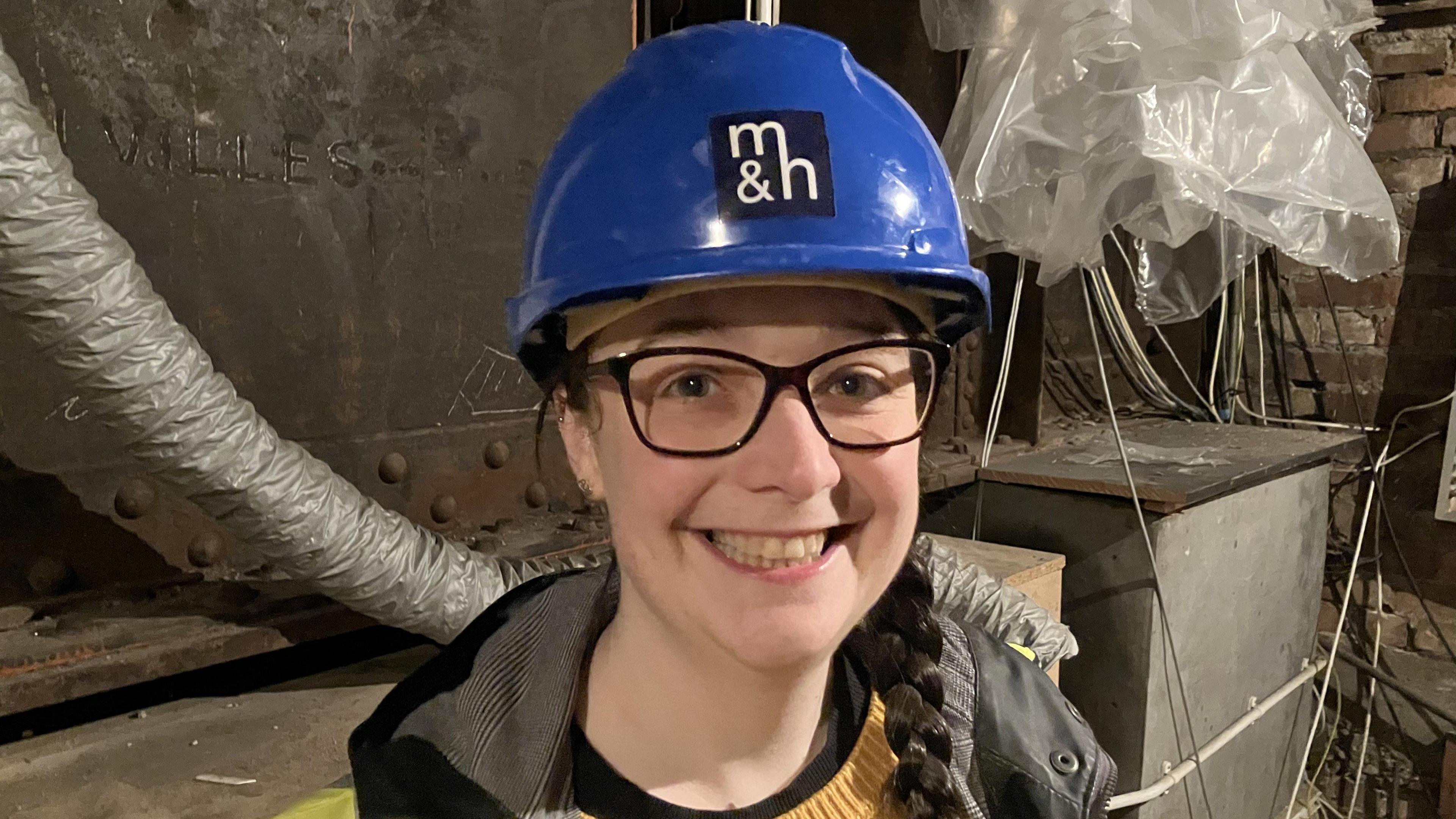 A woman with long dark brown hair in a plait is smiling and looking into the camera. She is wearing a blue hard hat, a yellow jumper and dark-rimmed glasses. 