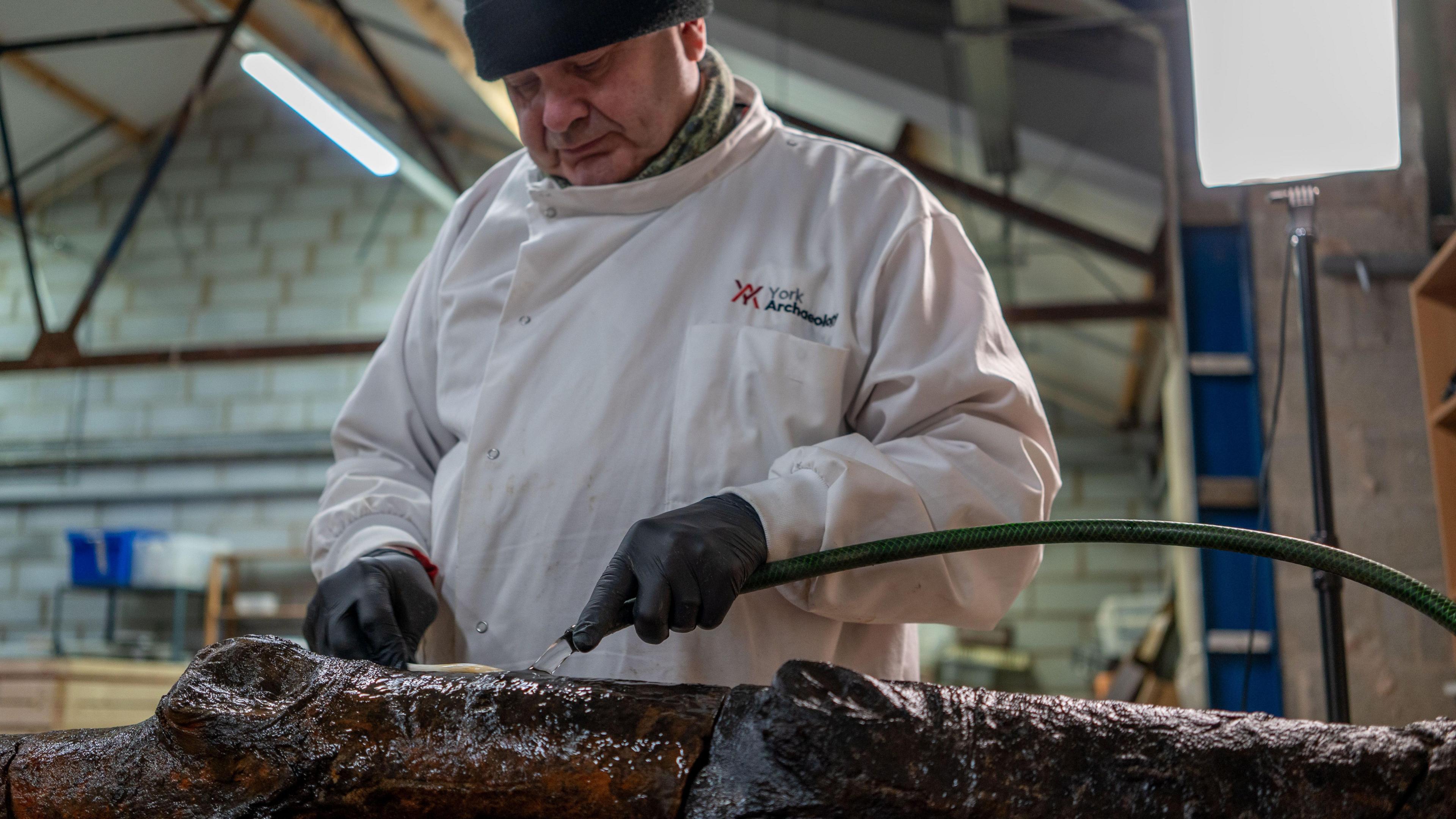 A man wearing a white laboratory jacket is pouring water on the wooden stake with a hose.