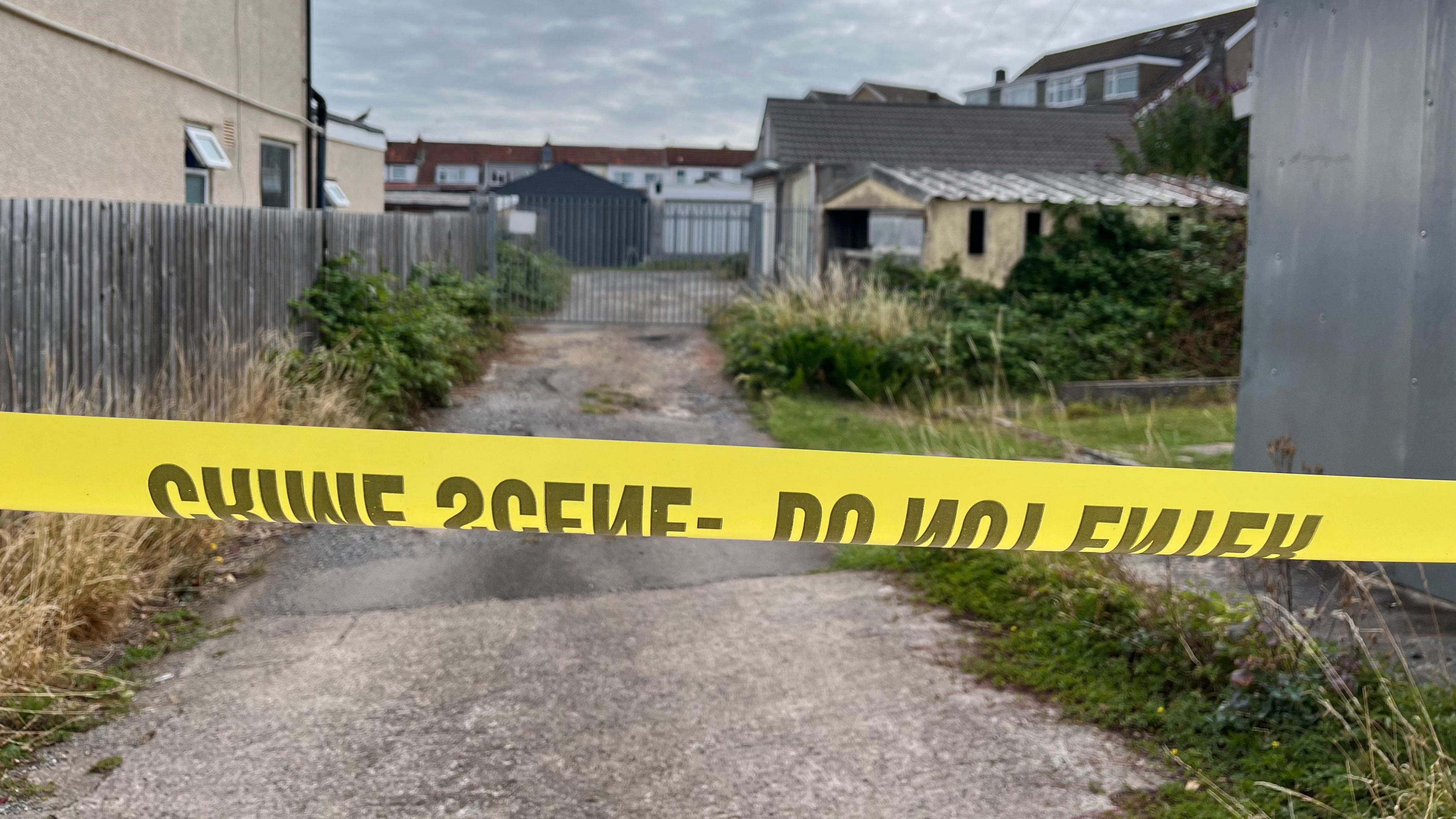 Yellow police tape closes off a lane with a metal gate halfway down - there isa run down garage on the right hand side and a property on the left hand side with moderate overgrowth running down either side of a central tarmacked roadway