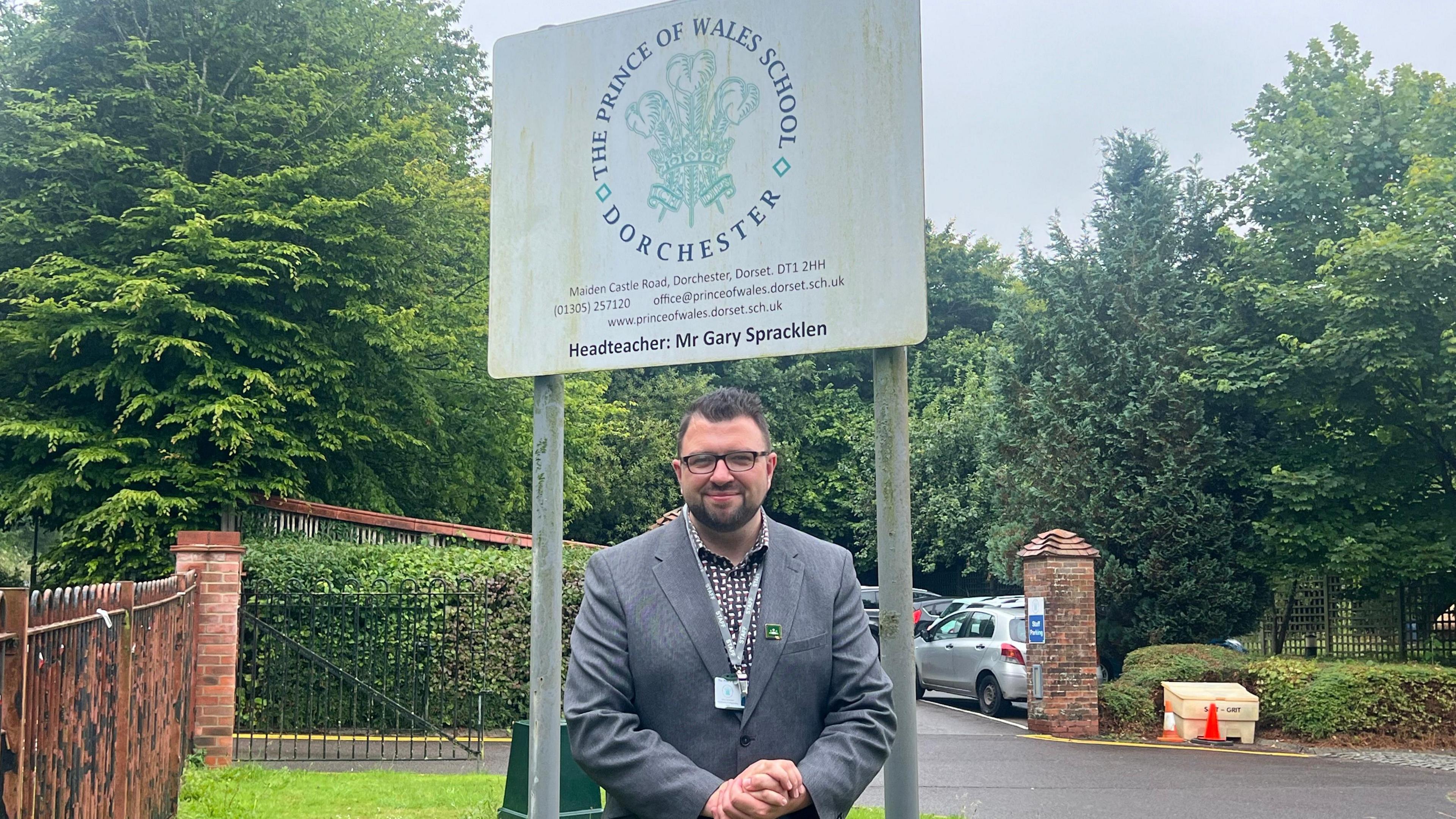 A man wearing a suit and glasses stands underneath a sign for his school, with a tree-lined car park behind him