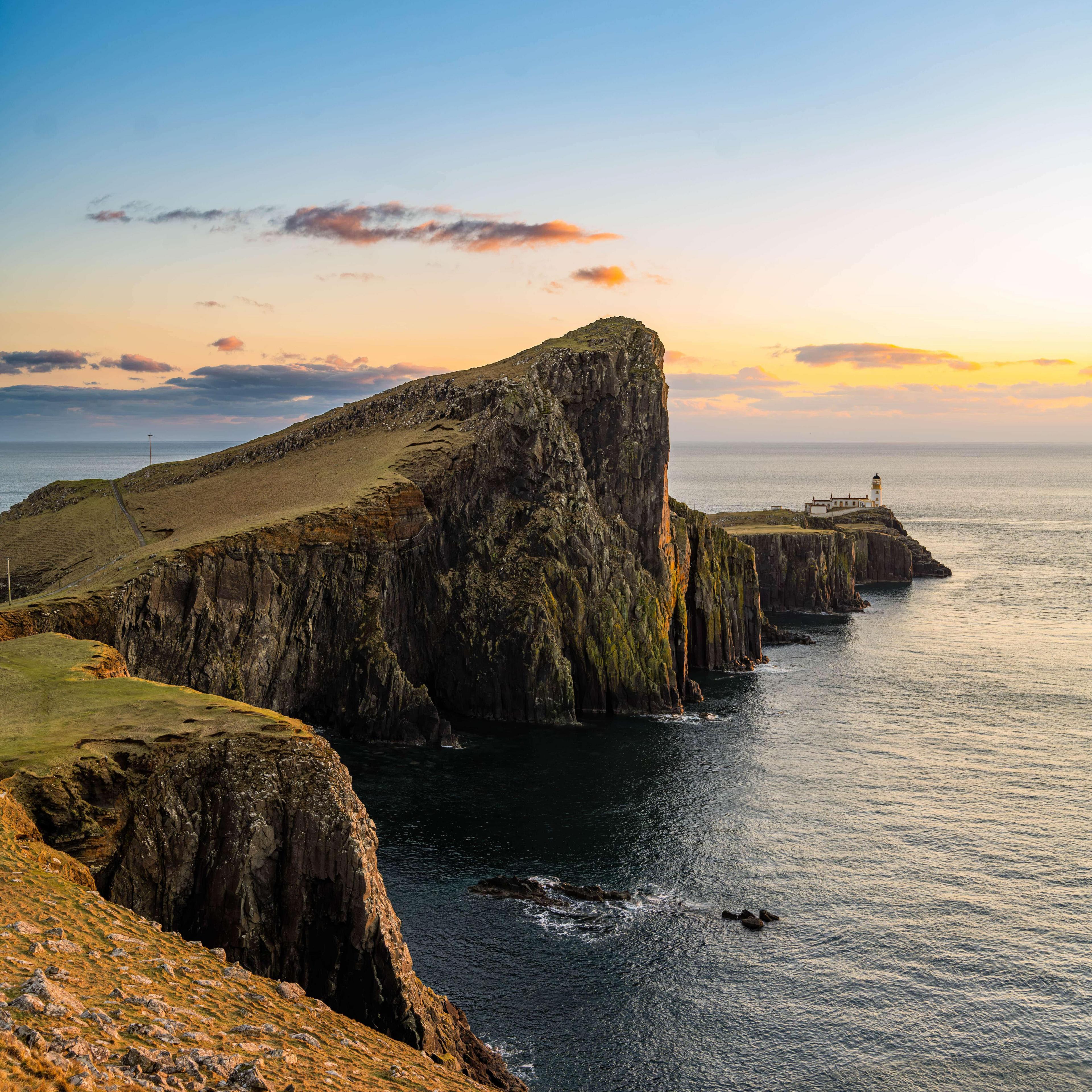 A view of Nest Point Lighthouse on Skye. The lighthouse can be seen in the background of the picture. In front of it are two large green ridges. The sky is orange and blue. The water below is also blue.