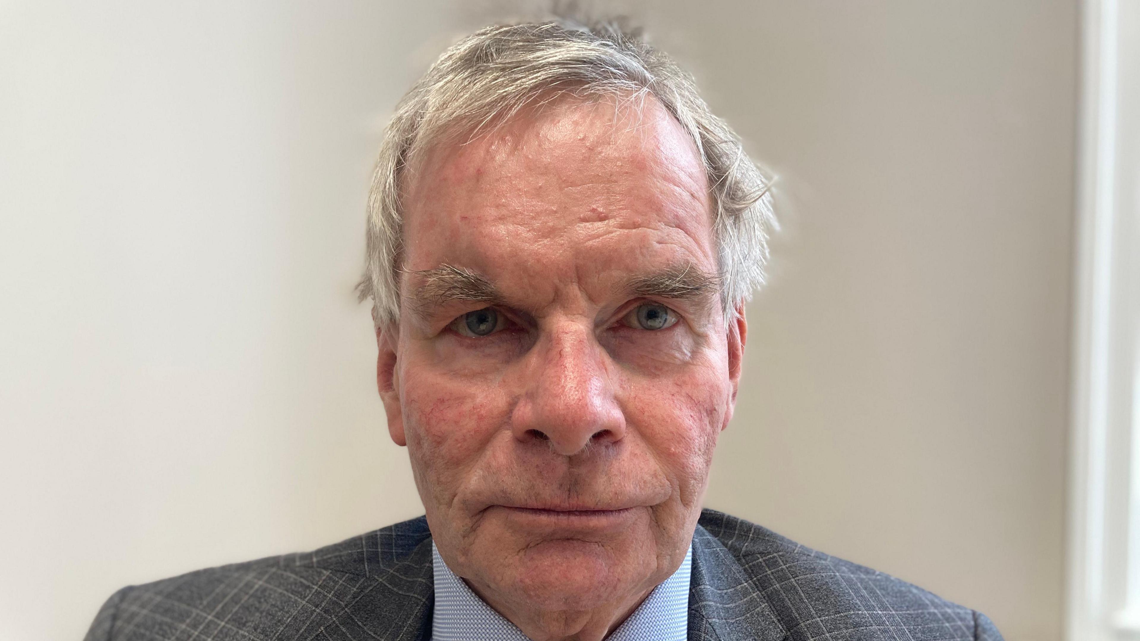 A close-up portrait of a man looking directly into the camera with a serious expression. He has short grey hair and is wearing a grey blazer and light blue shirt. The wall in the background is white.