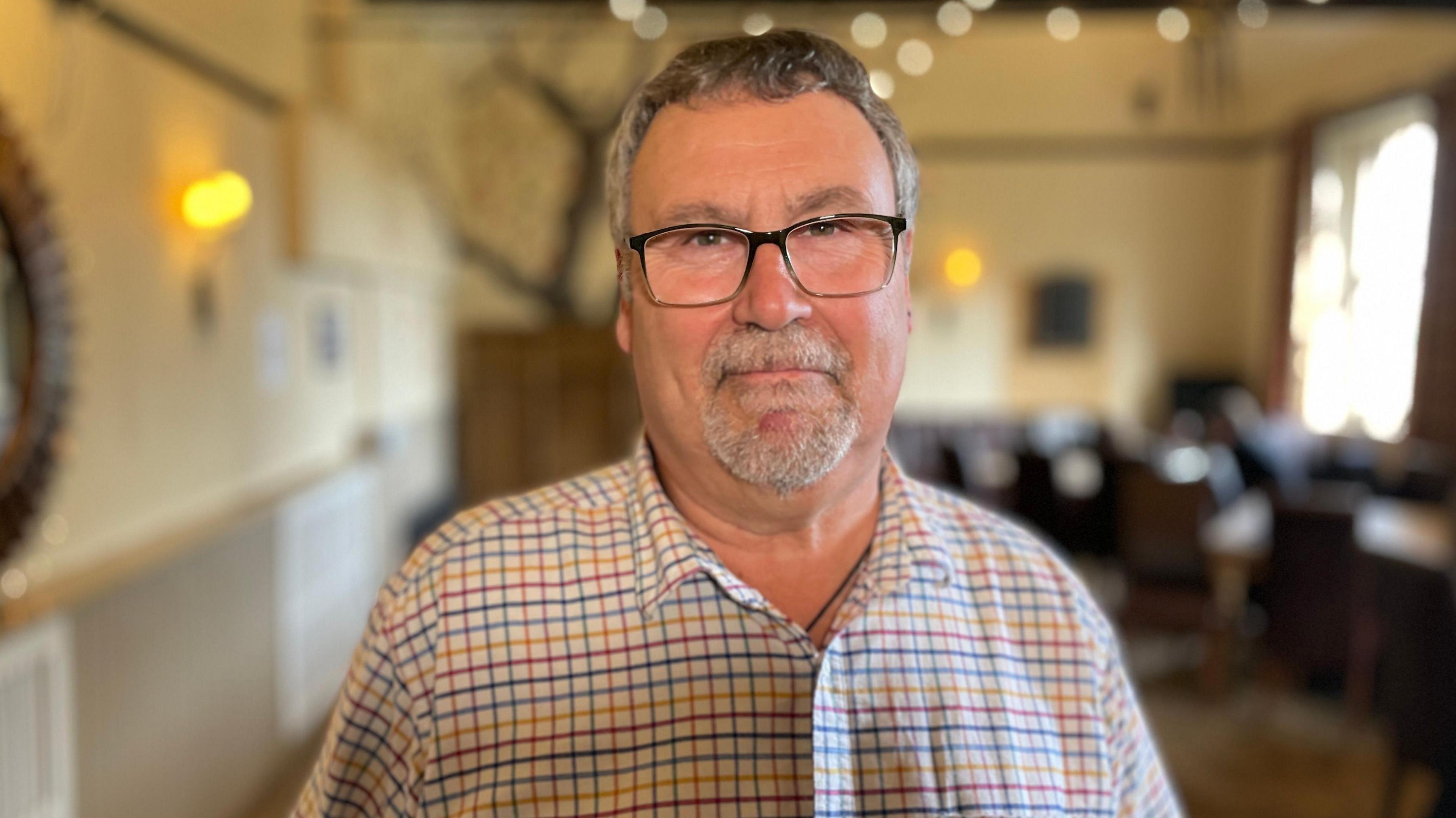 Nigel Gisby wearing a colourful checked shirt and black framed rectangular glasses. He has black and grey hair and facial hair. He is standing inside the pub, which has cream walls, wooden furniture and warm overhead lighting.