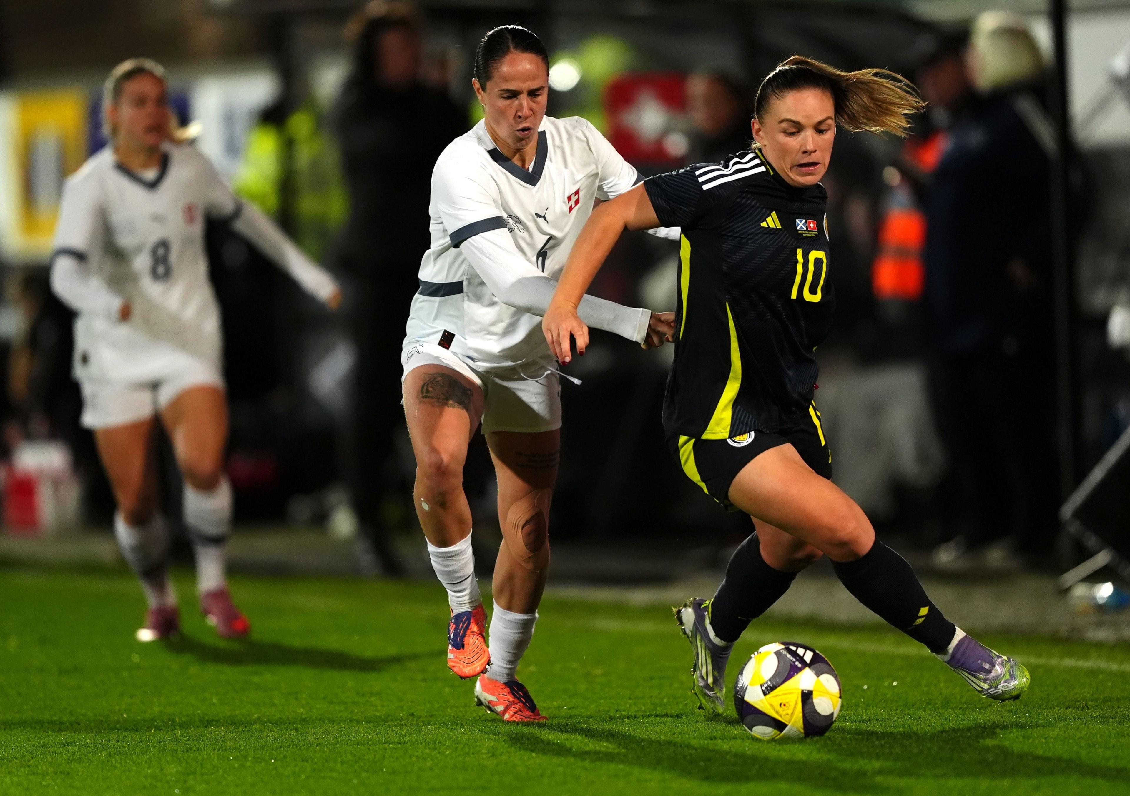Switzerland's Geraldine Reuteler (left) and Scotland's Kirsty Hanson battle for the ball during. Geraldine wears a white kit with dark blue accents while Kirsty wears a black shorts and top combo with yellow accents and a yellow number 10.