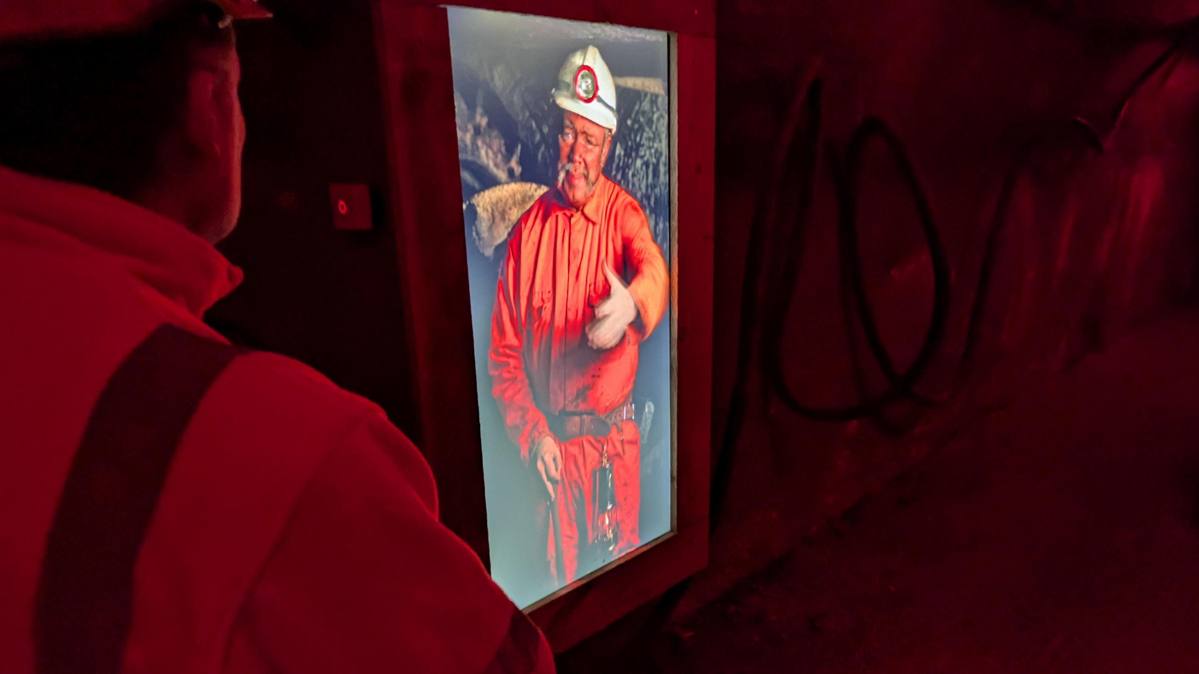 An animated digital billboard of a mineworker in a helmet. The billboard is in a dark tunnel.