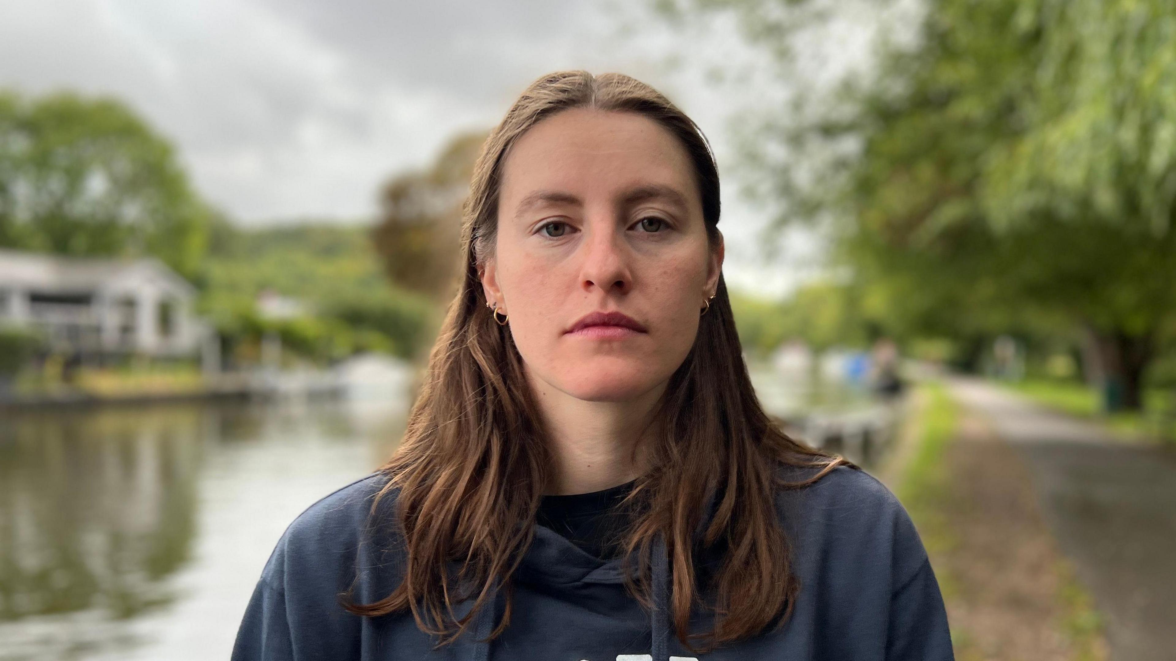 Amber has long brown hair. She is looking at the camera with a serious look on her face, standing by the river.