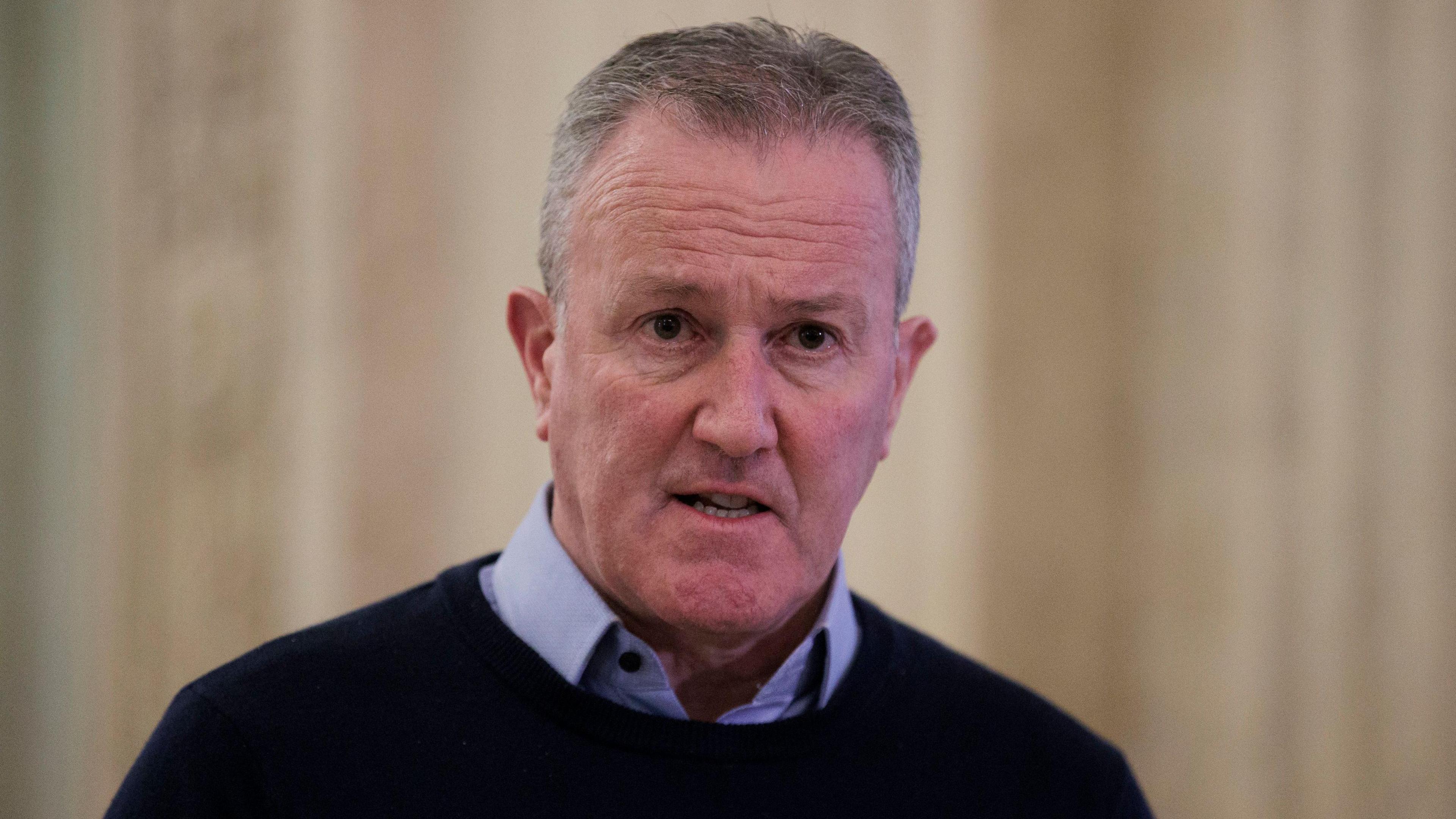 Conor Murphy - a man with short grey hair wears a dark jumper over the top of a blue collared shirt. He is standing in the main hall at Stormont, but the background is blurred.