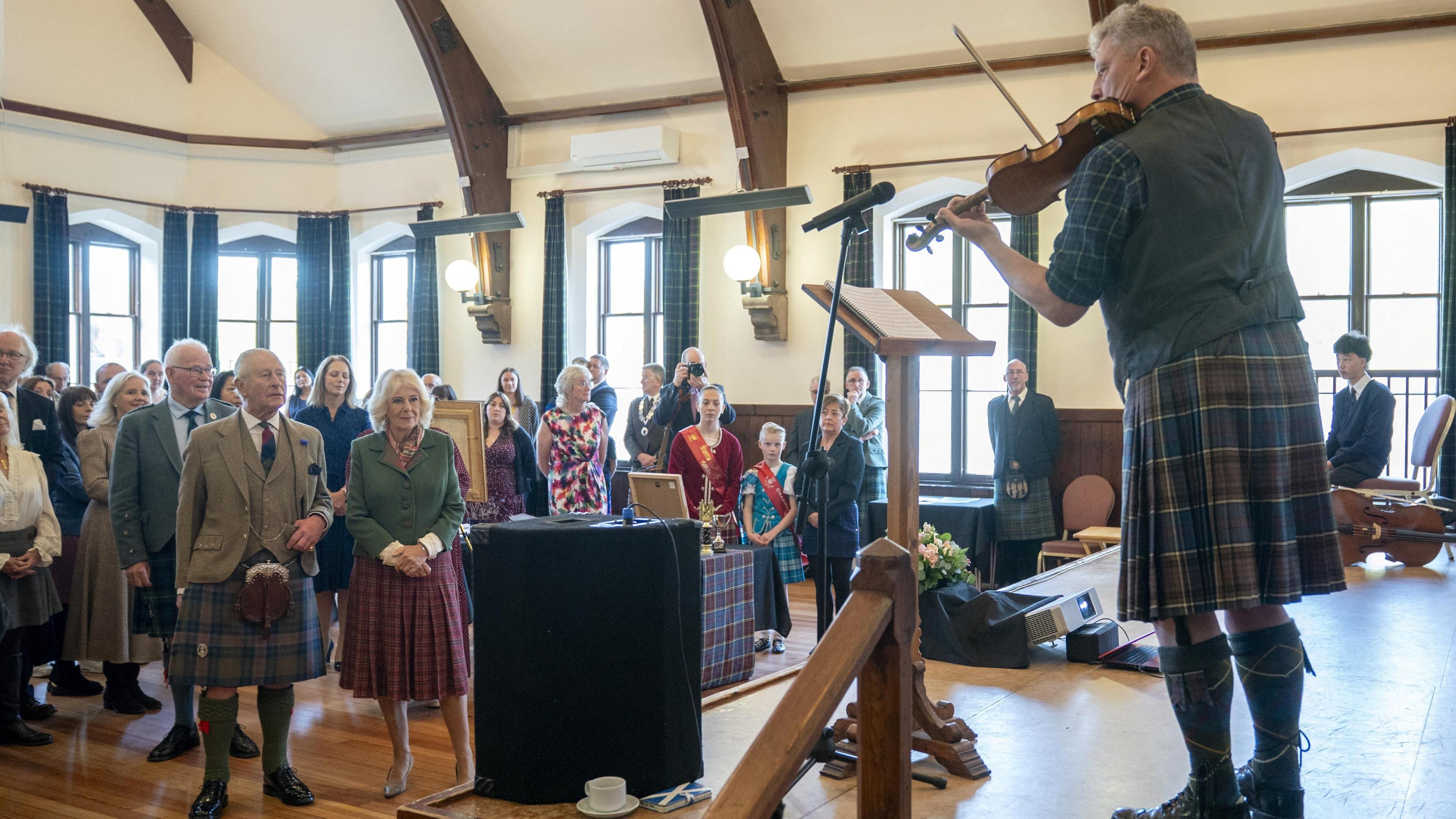 King Charles III and Queen Camilla are among a crowd watching the first performance of a fiddle tune composed by local musician Dr Paul Anderson, who is playing on a stage, during a visit to mark the 150th anniversary of The Albert Hall