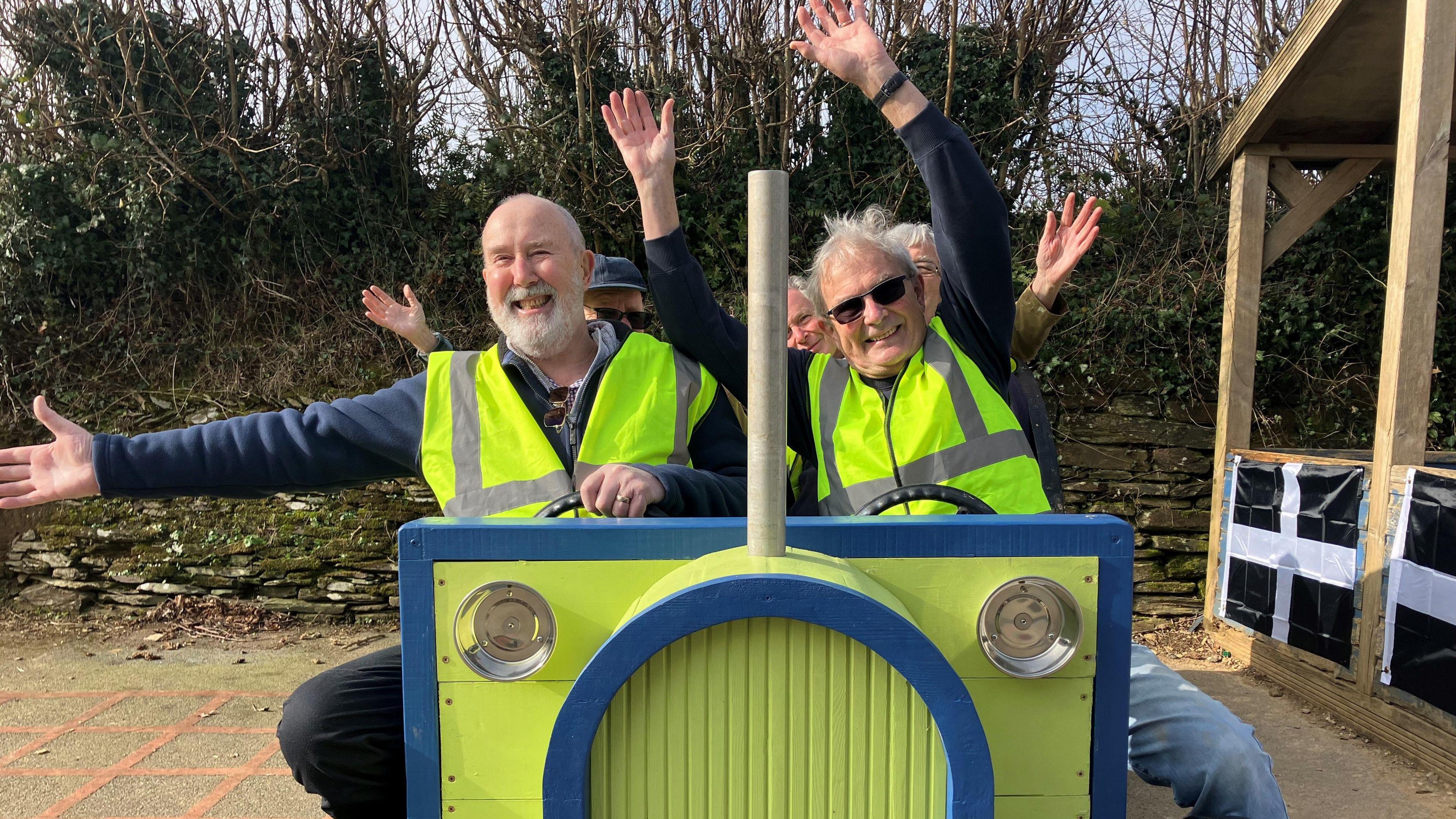 A group of men wearing high viz jackets and waving their hands in the air as if they are flying left on the wooden tractor. 