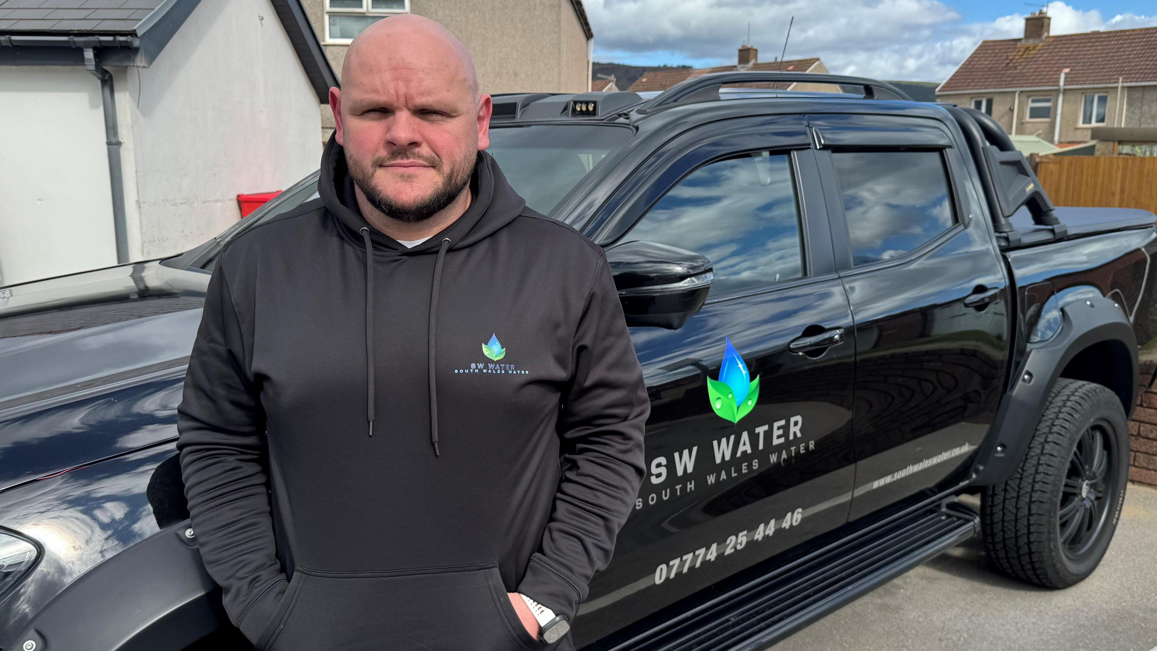 A bald man wearing a black hoodie branded “South Wales Water” stands in front of a black pickup truck with matching company logos and a phone number printed on the side. Behind him are residential houses under a partly cloudy sky.